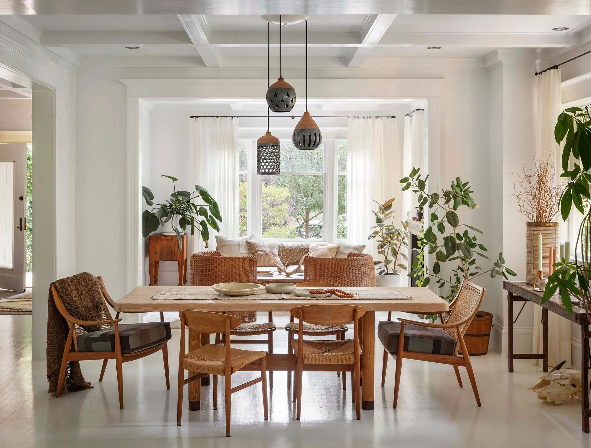 A light wood dining table with woven cane chairs stands on white painted floors under three pendant lights, with a living area visible behind it.