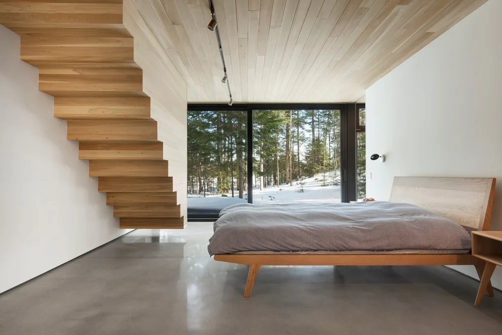 A minimalist bedroom features a floating wood staircase, a wooden bed, polished concrete floors, and a glass window overlooking a snowy forest.