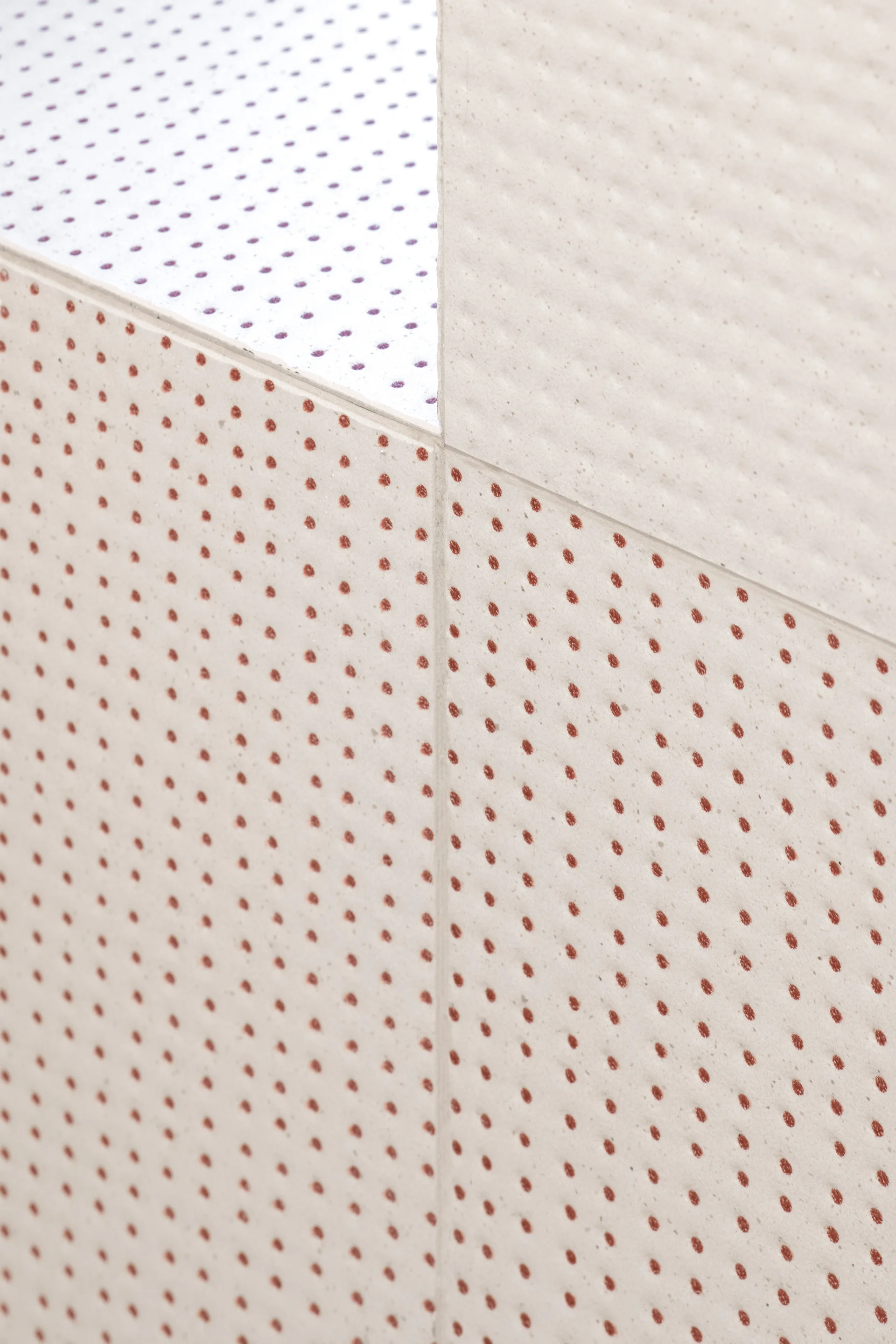 A close-up view of textured off-white ceramic tiles with a repeating pattern of small terracotta and purple polka dots separated by a thin grout line.