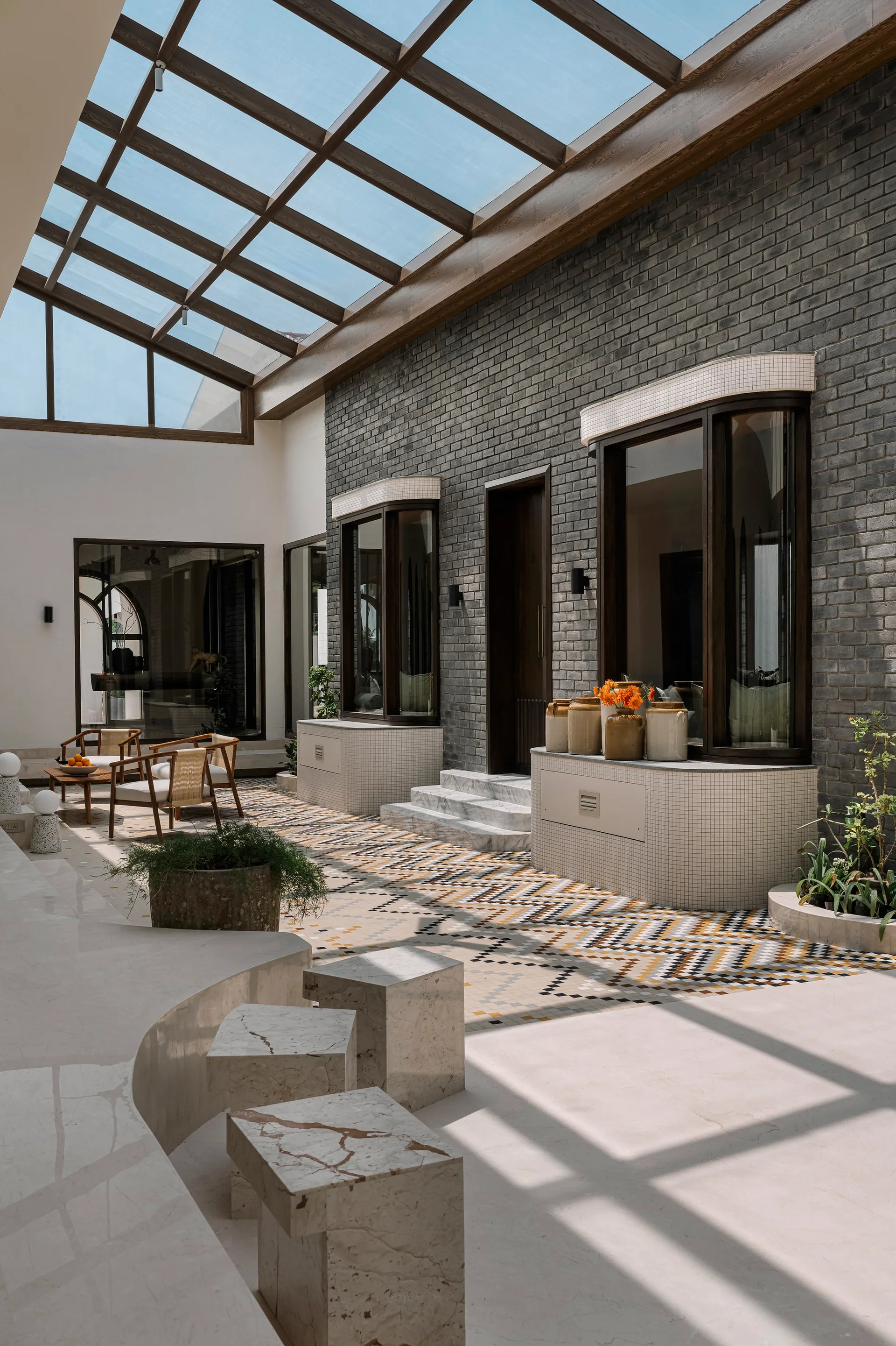 An airy atrium courtyard features a glass skylight roof, a grey brick wall, and colorful geometric patterned floor tiles.