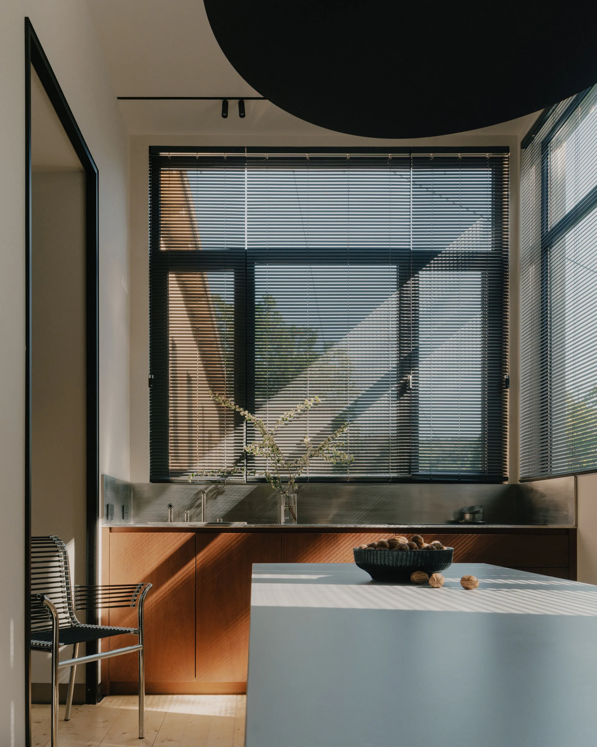 A modern kitchen featuring warm wood base cabinets, a brushed metal backsplash, a white island, and sunlight filtering through slatted window blinds.