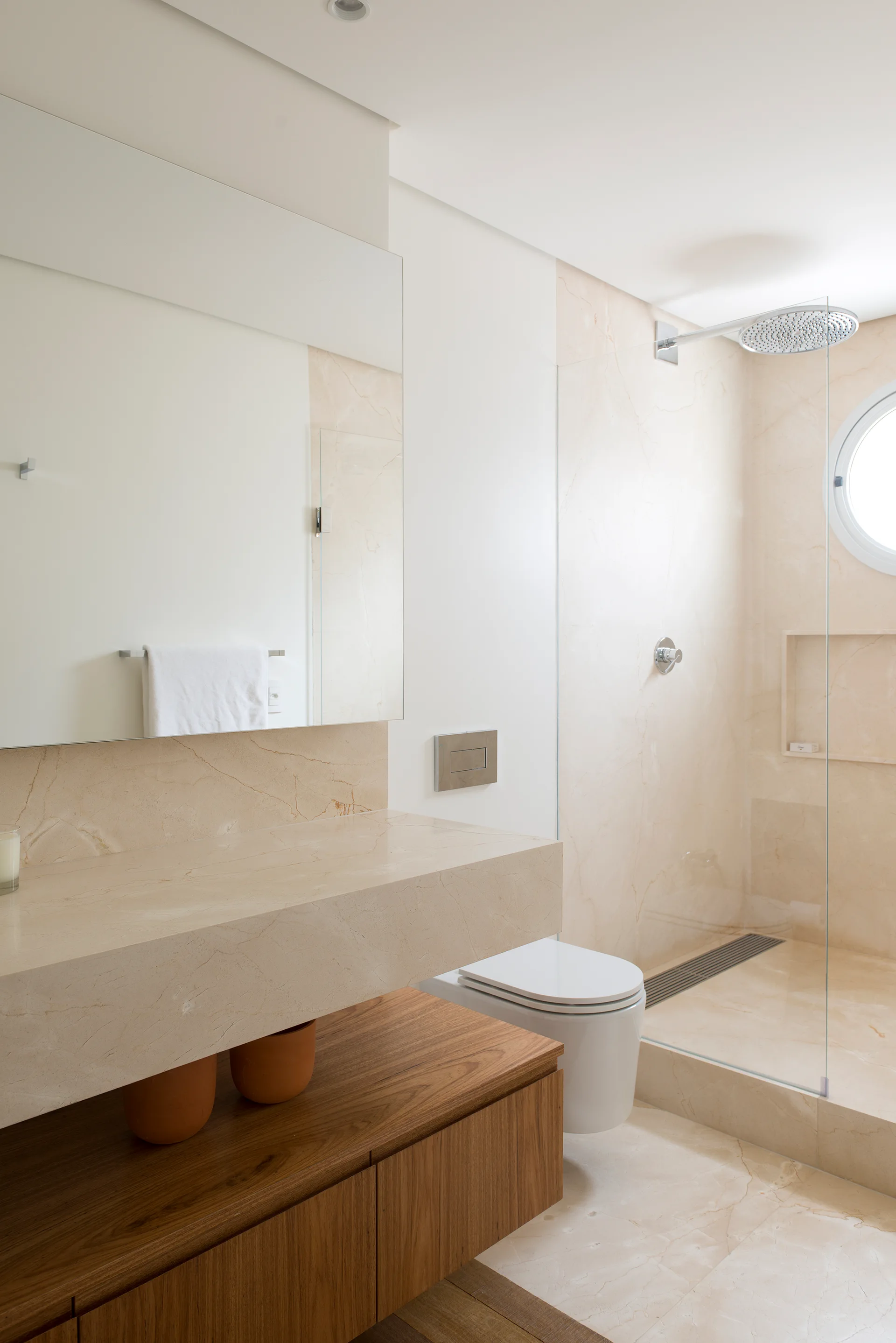 A modern bathroom featuring veined beige marble walls, a thick floating stone vanity above a wood cabinet, a wall-mounted toilet, and a glass shower.