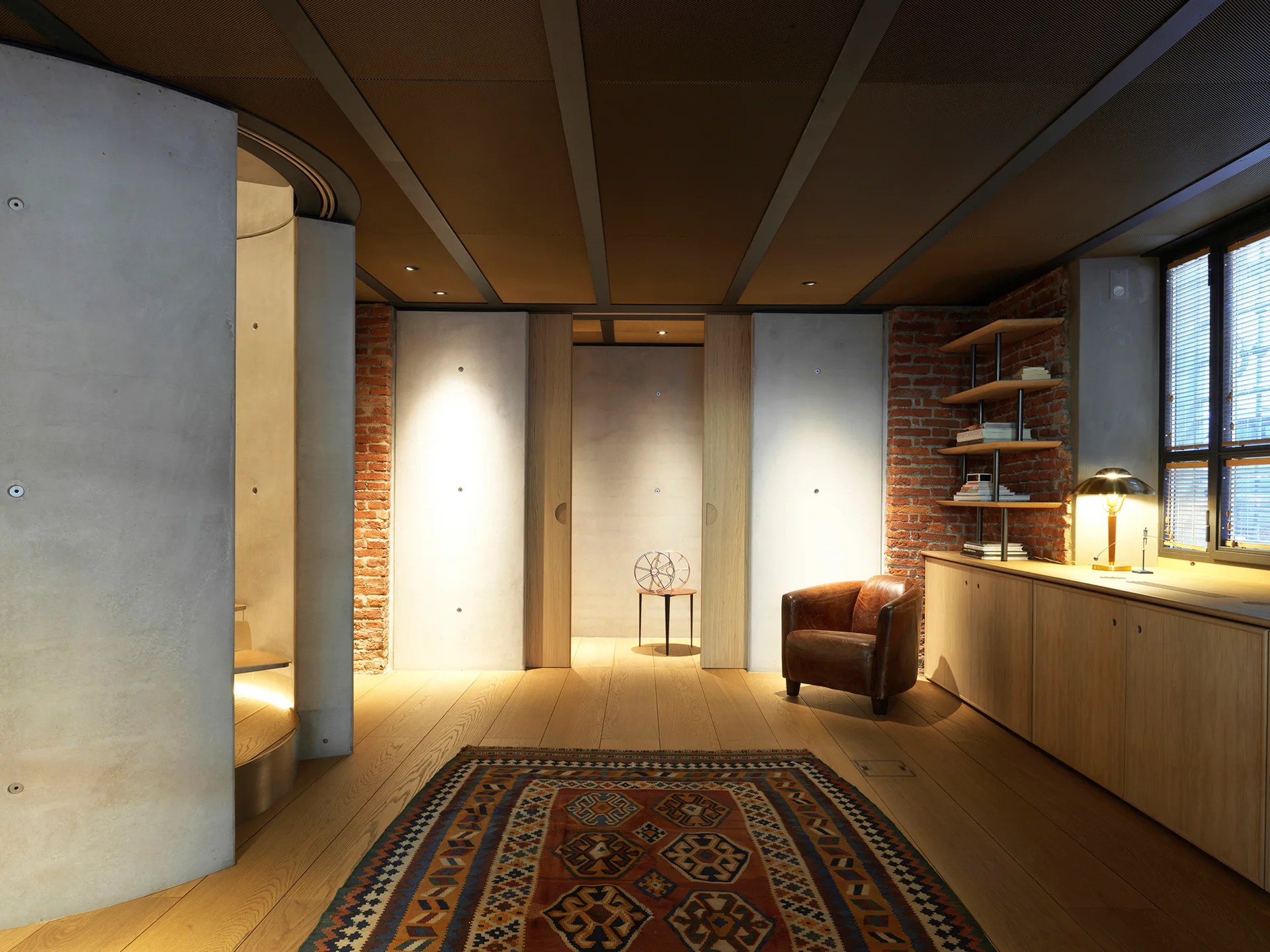 An industrial lounge area features smooth concrete and exposed brick walls, a leather armchair, a patterned rug, and a light wood cabinet.