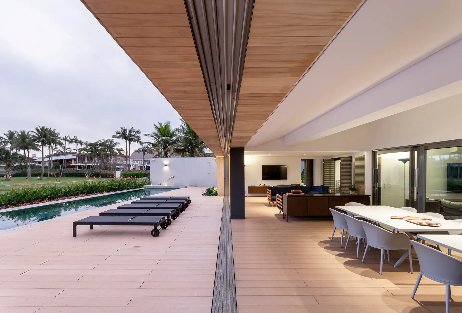 A modern covered patio features a wood-plank deck with black lounge chairs next to a swimming pool, an outdoor dining table, and large sliding doors.
