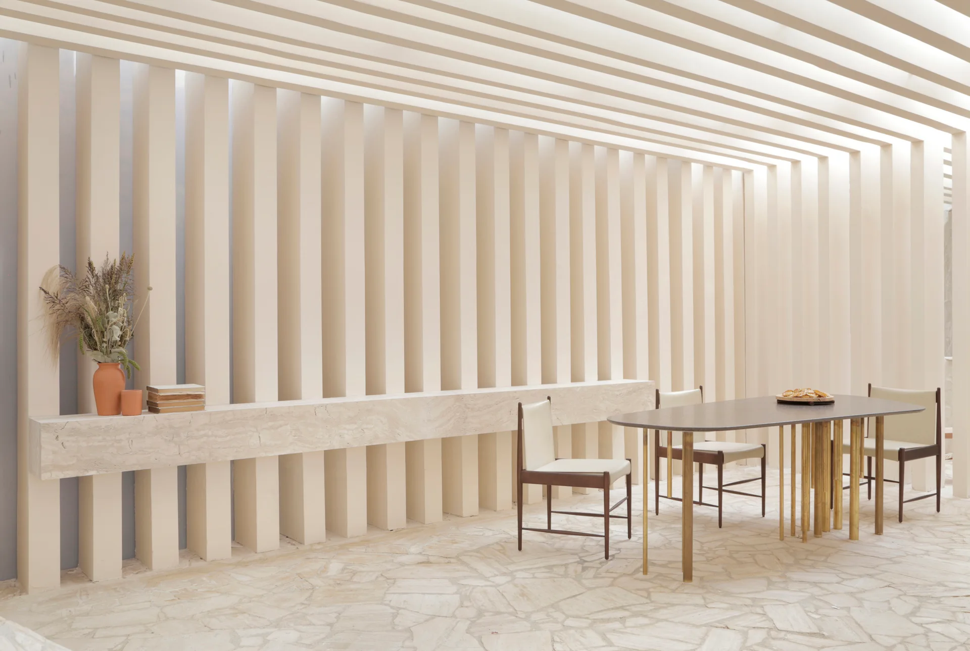 A minimalist dining room features a dark oval table and chairs set against a textured beige slat wall with an intersecting stone ledge.
