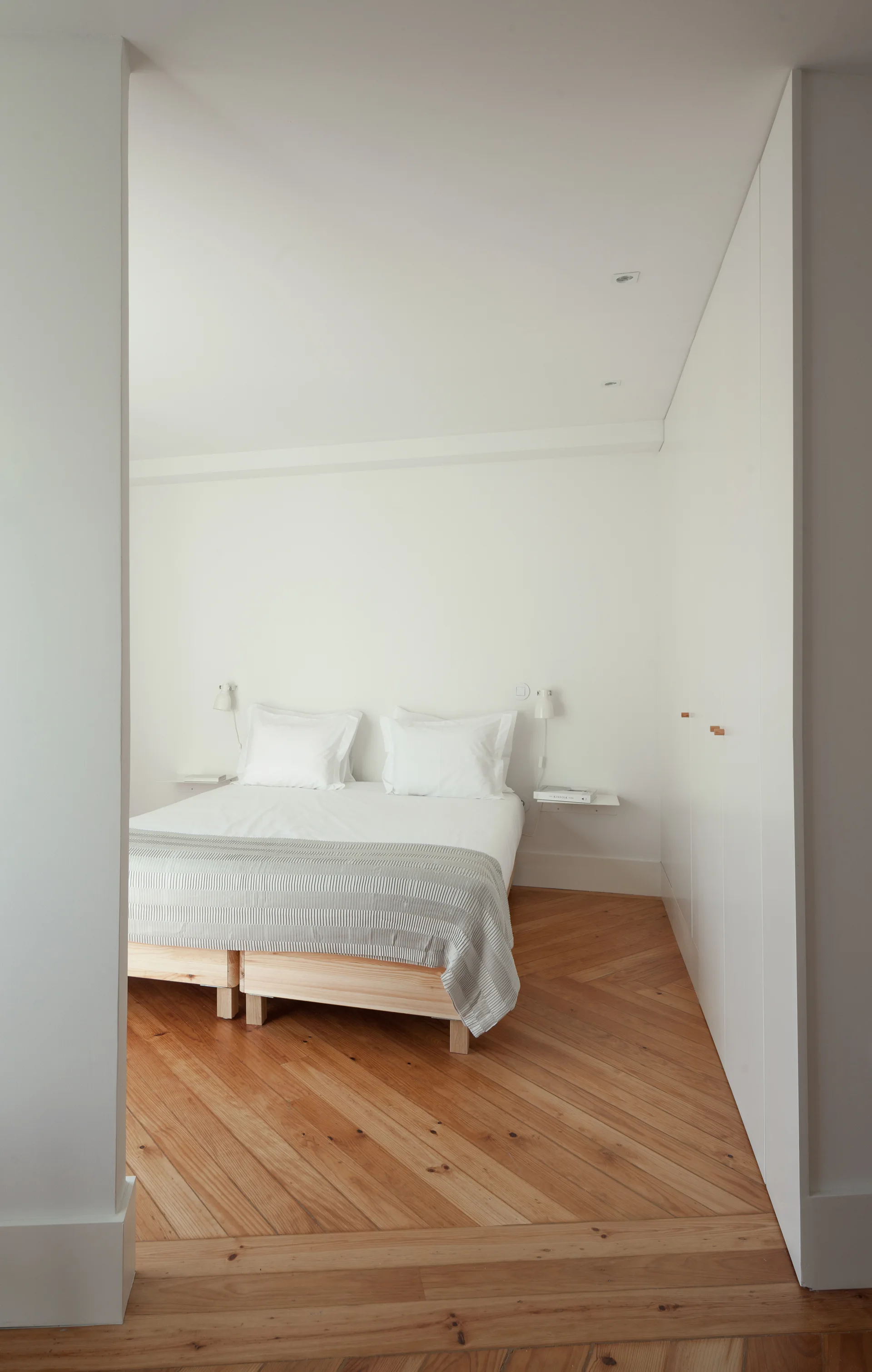A minimalist white bedroom featuring a light wood bed frame with white linens on chevron pattern wood floors next to built-in closet panels.