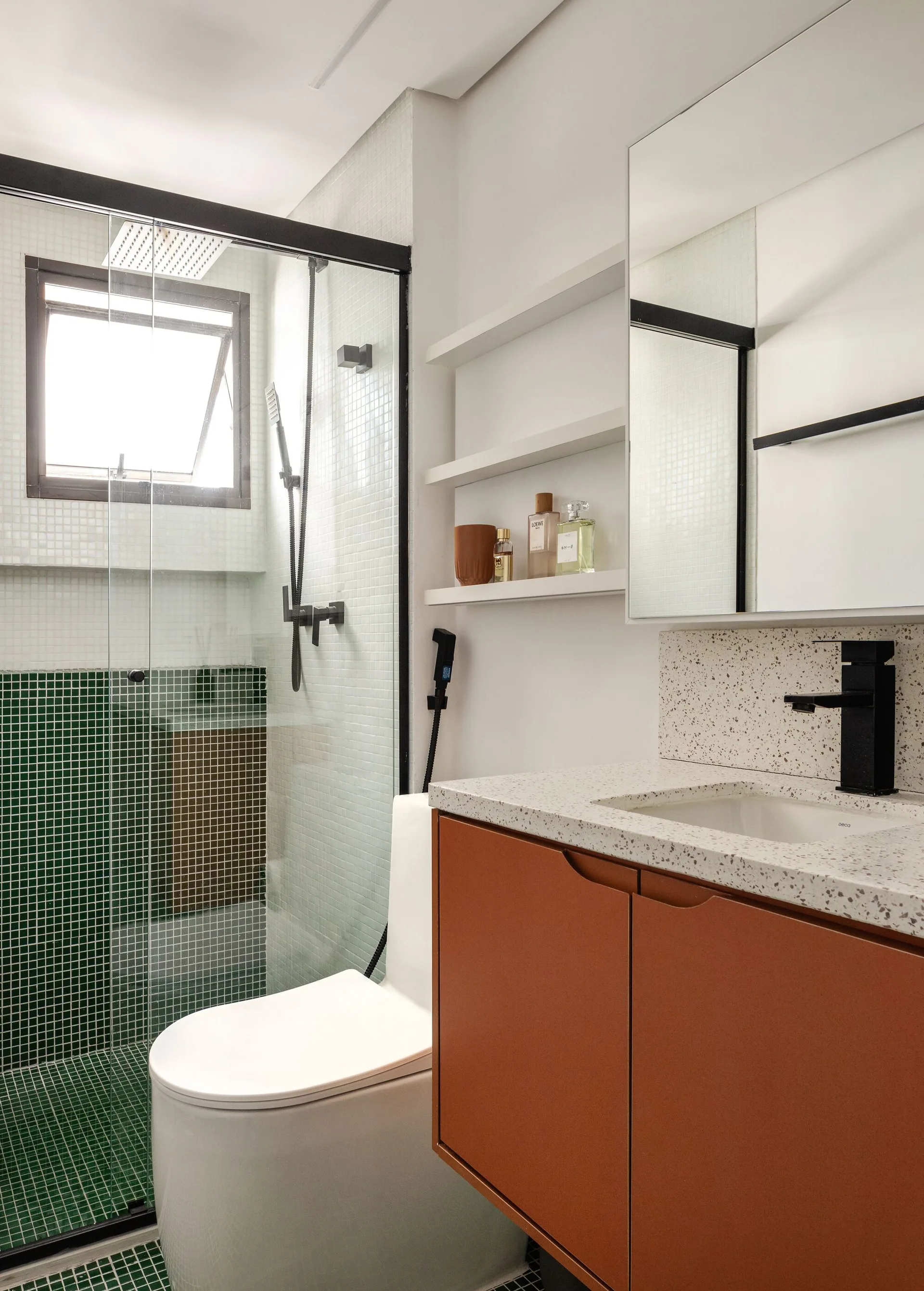 A modern bathroom features a green mosaic-tiled shower, a white toilet, and a terracotta orange vanity with a speckled countertop and black faucet.