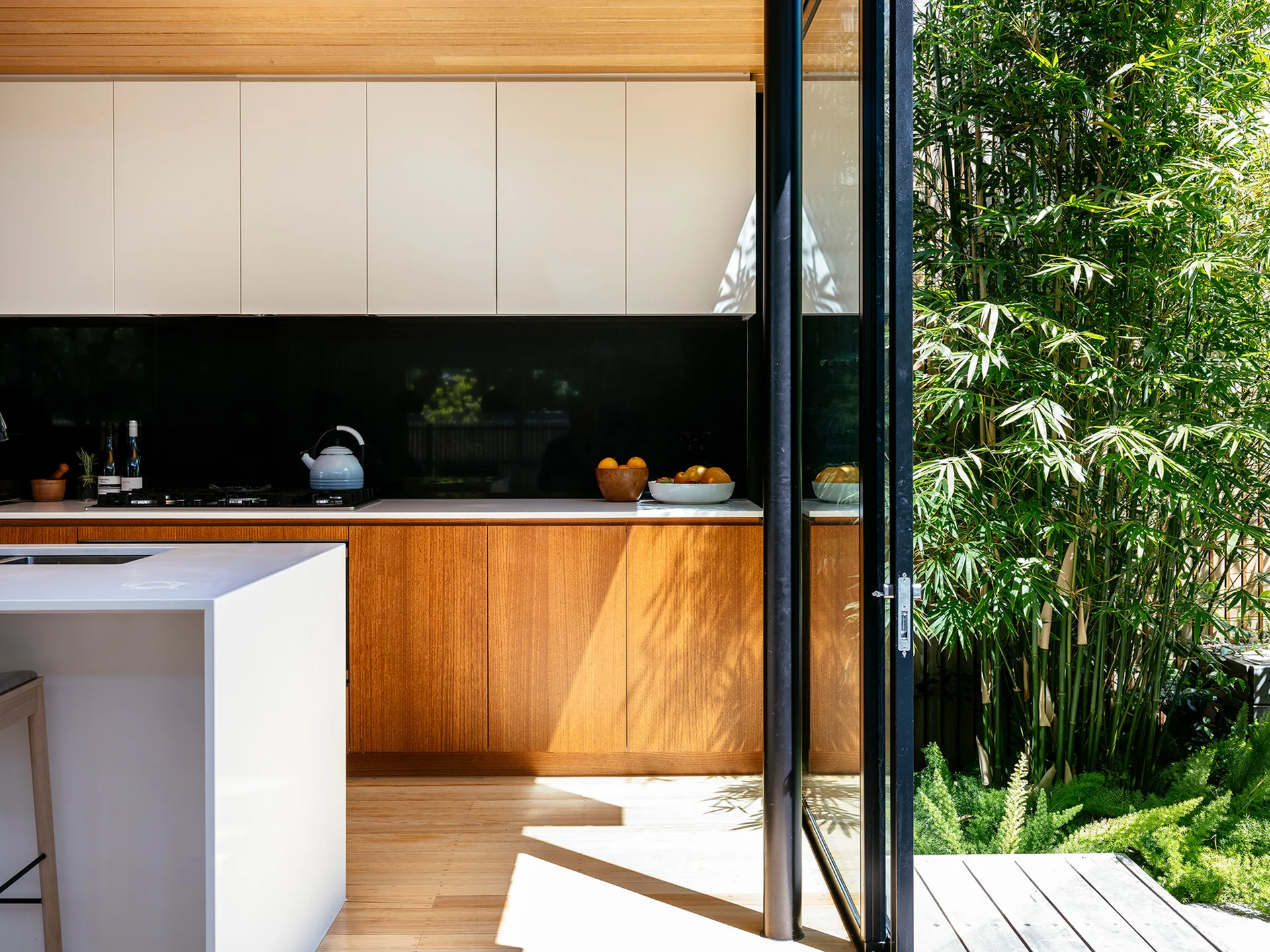 A modern kitchen with wood and white cabinets features an open glass door that leads to an outdoor patio with tall green bamboo.