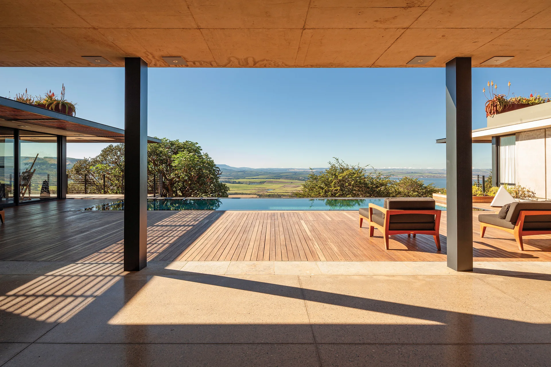 A modern covered terrace with concrete ceiling, dark columns, a wooden deck, lounge chairs, and an infinity pool overlooking an expansive landscape.