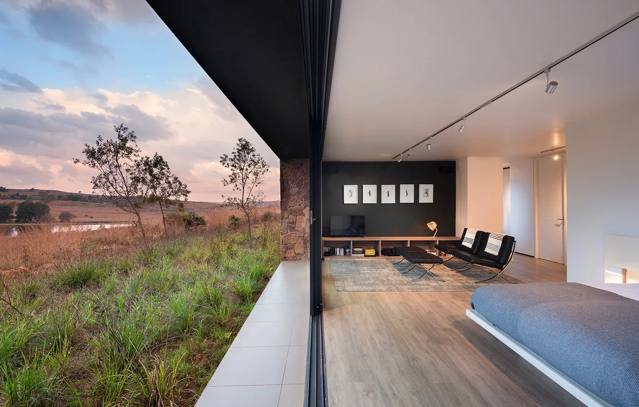 A modern bedroom features a large retractable glass door opening to a grassy outdoor landscape, with a low-profile bed and black leather seating.