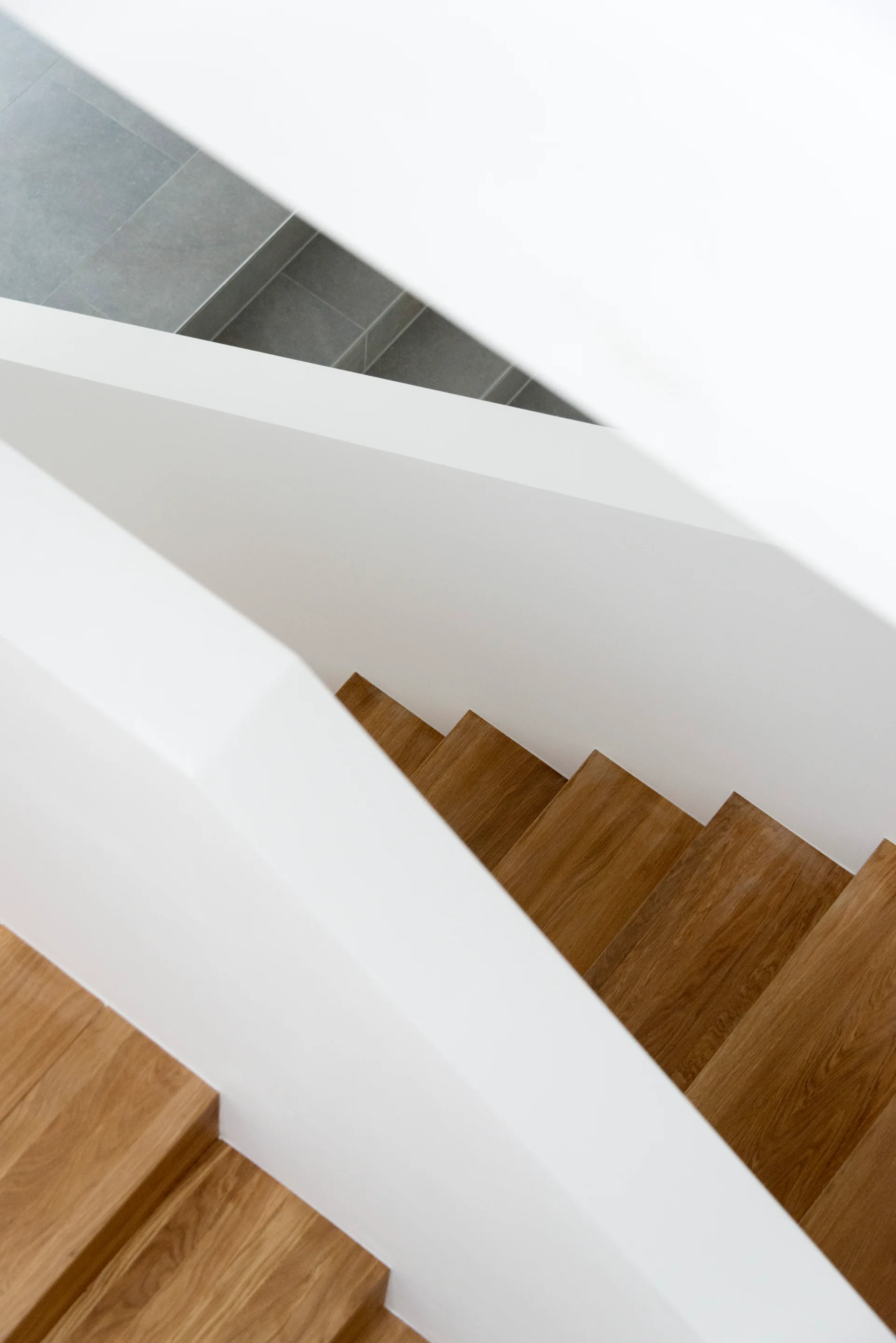 A high-angle view of a minimalist modern staircase featuring warm wood treads nestled within sculptural, solid white walls and a gray tiled landing.