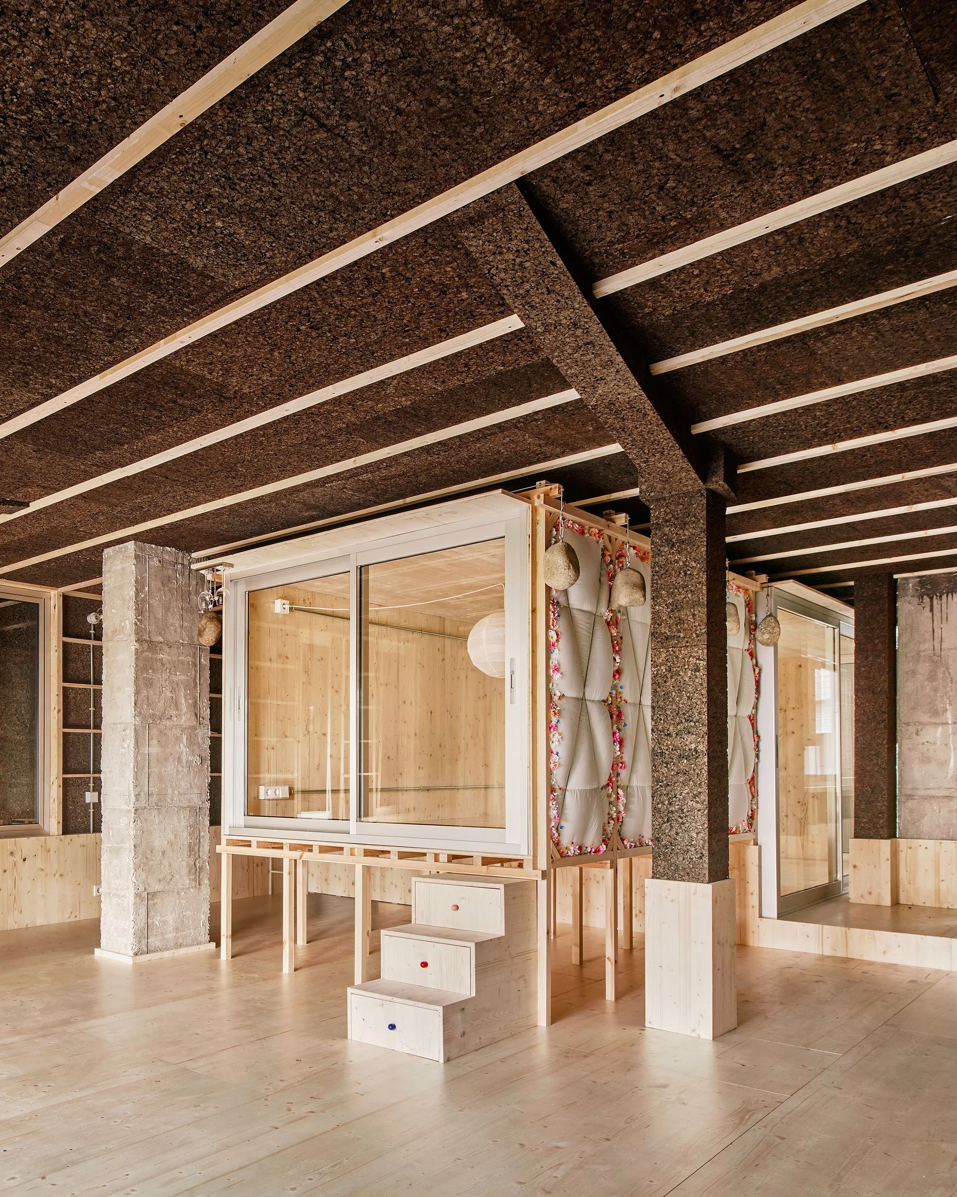An elevated wooden cabin structure stands inside a room featuring a textured dark cork ceiling with light timber beams and a concrete pillar.