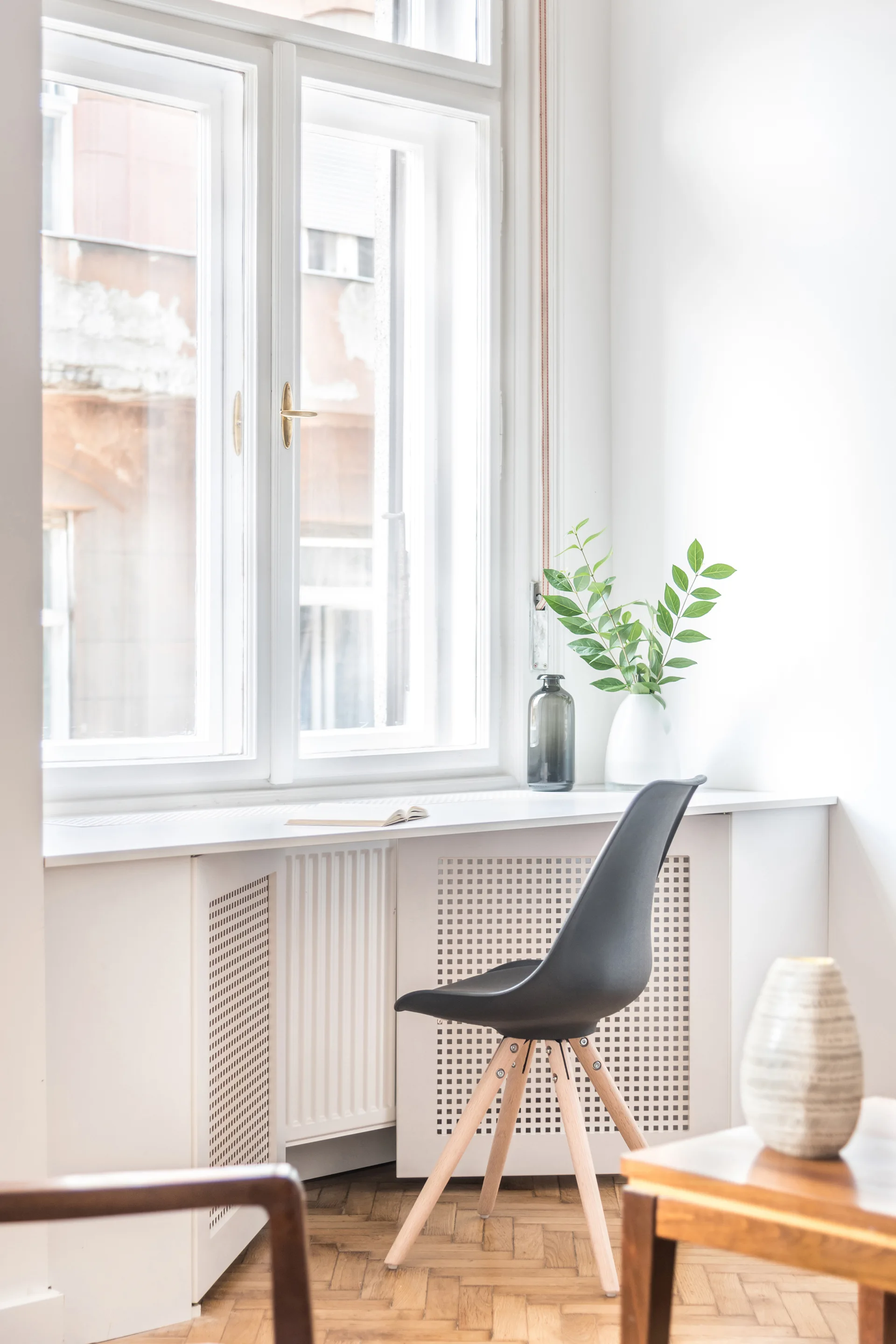 A black chair sits at a white built-in desk under a large window with perforated radiator covers and herringbone parquet flooring.