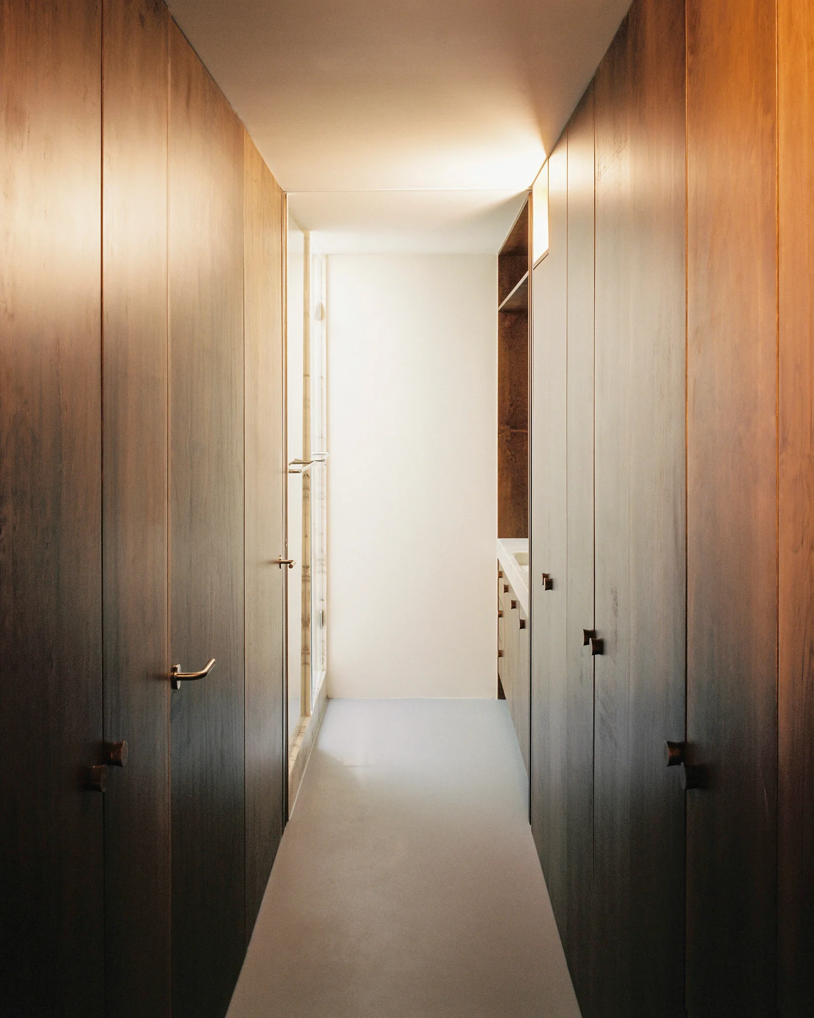 A narrow minimalist hallway lined with floor-to-ceiling dark wood paneled storage cabinets, a light stone floor, and a glowing ceiling cove.