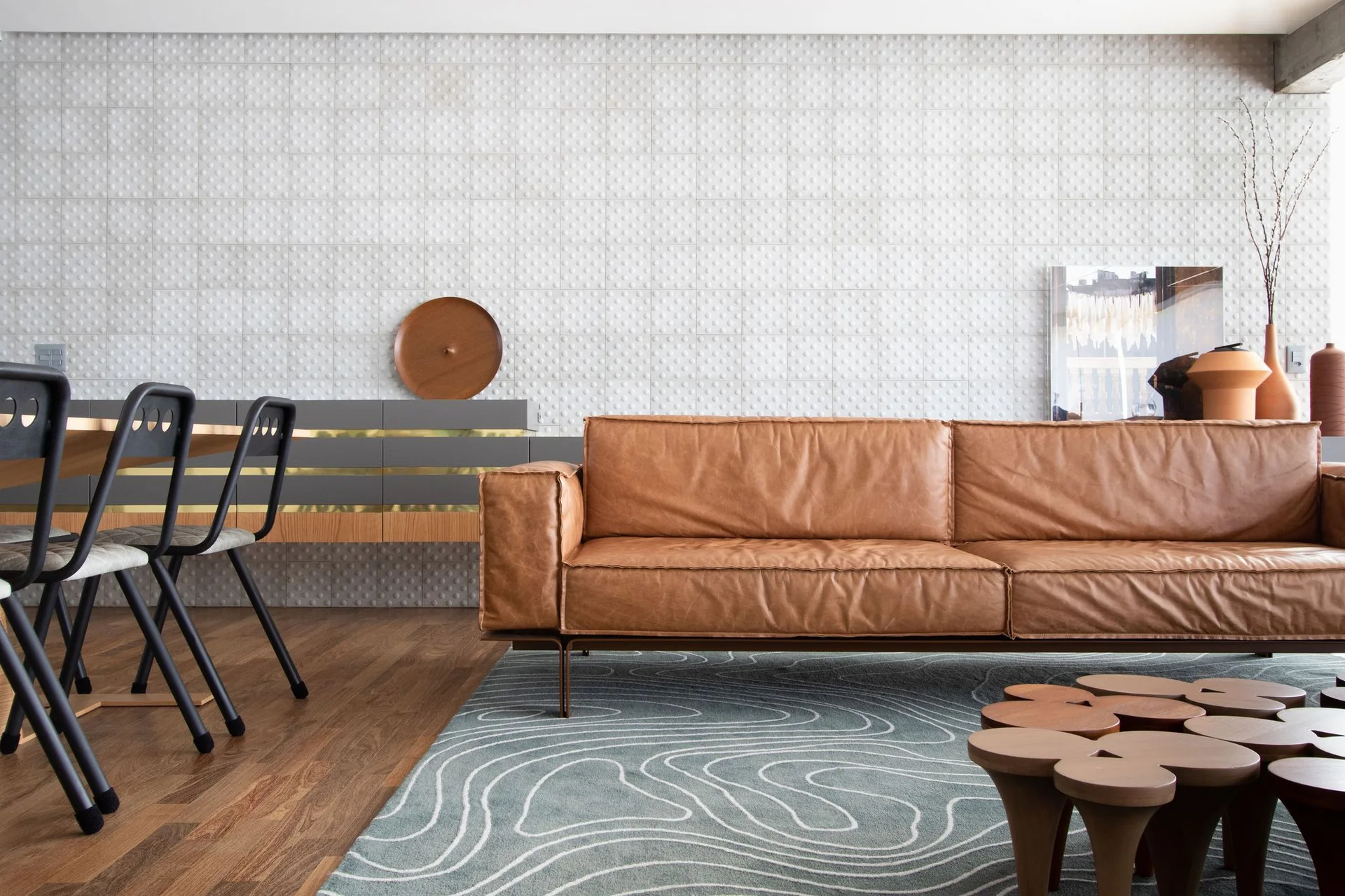 A modern living room features a tan leather sofa, a topographic pattern rug, and a textured white grid accent wall behind a low grey sideboard.