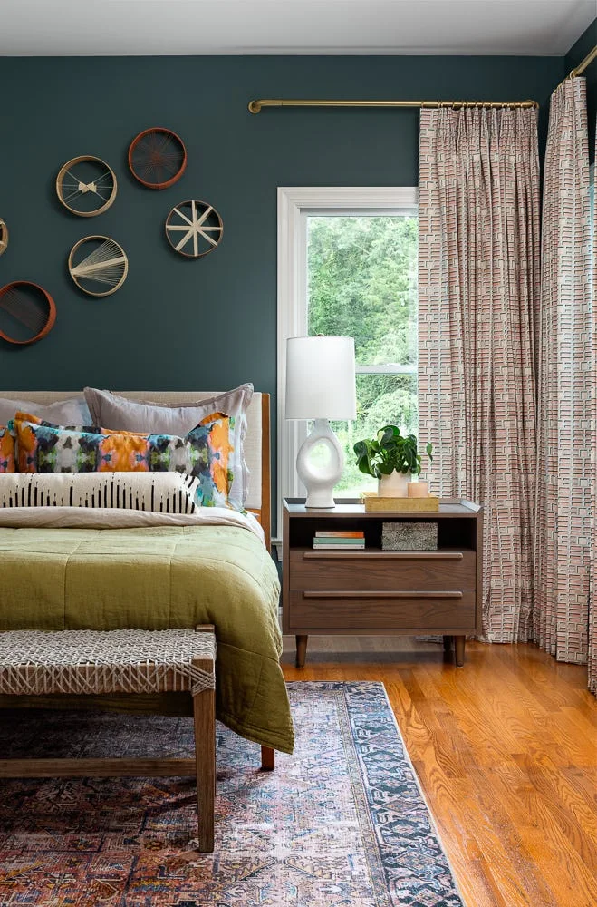 A bedroom with a dark green accent wall, wooden nightstand, a white ceramic lamp, and patterned curtains hanging from a brass rod.