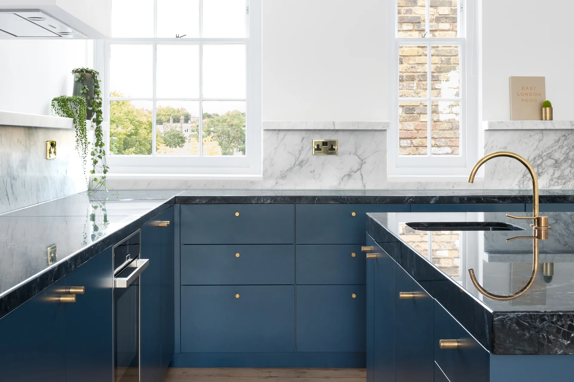 A kitchen with matte blue cabinetry, dark stone countertops, a brass faucet, and white marble backsplash pieces under large sash windows.