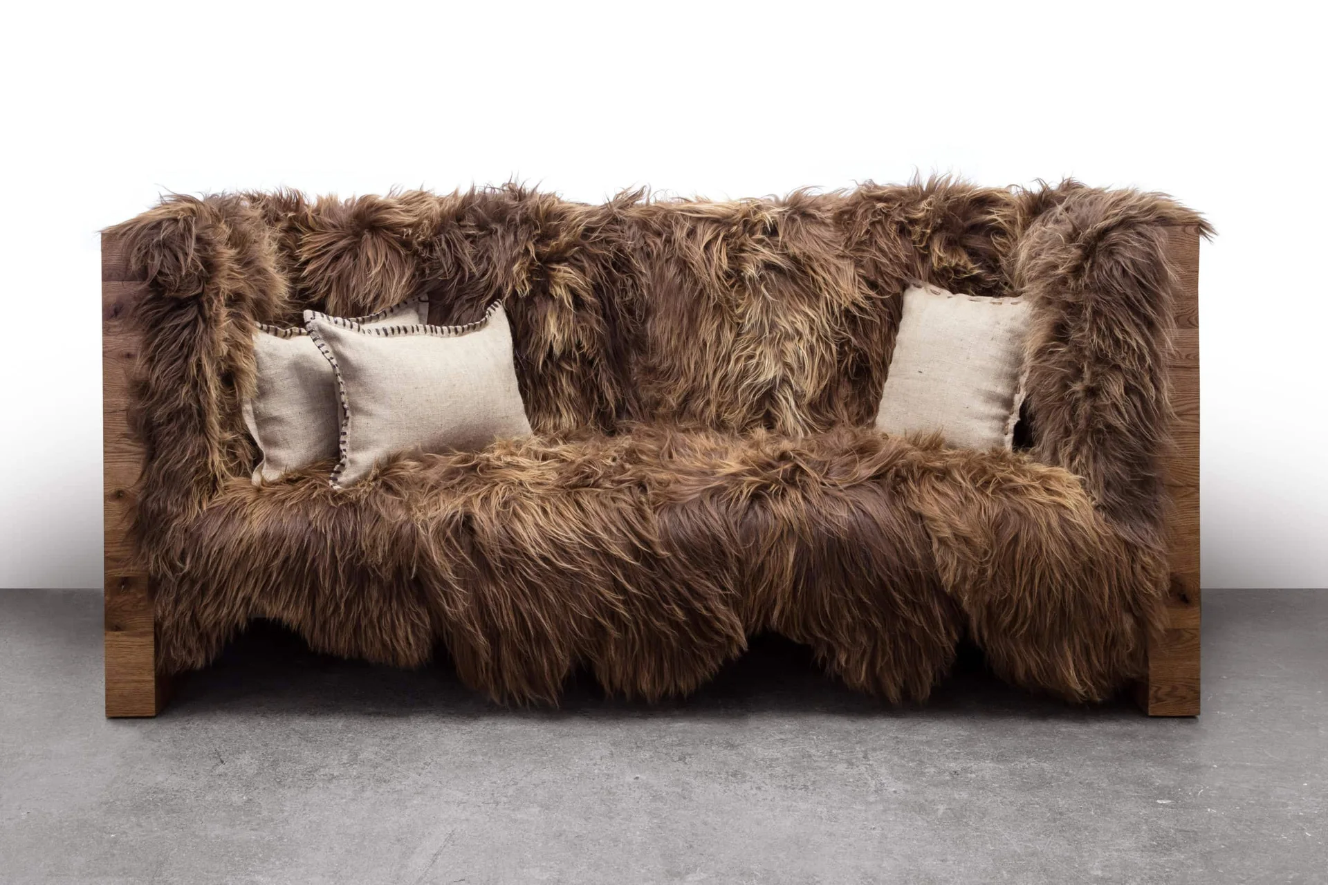 A rustic wooden frame sofa covered in shaggy brown fur with three beige linen throw pillows on a gray concrete floor.