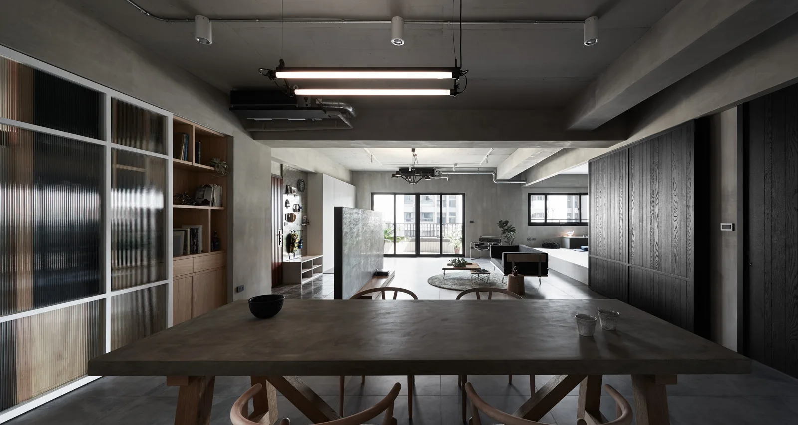 An industrial dining area features a large concrete dining table under linear light fixtures next to a ribbed glass partition.