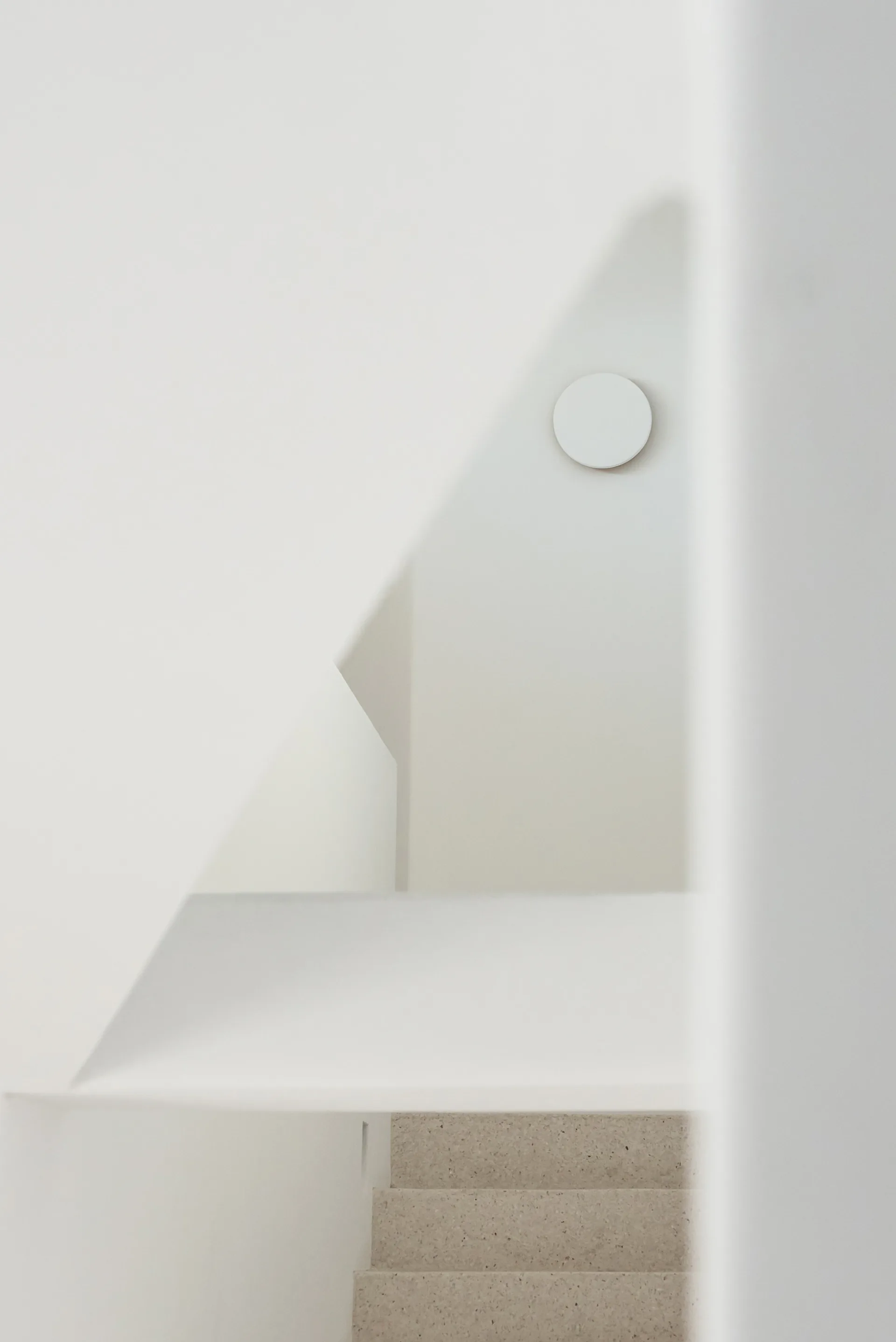 A minimalist white stairwell featuring geometric plaster walls, textured beige stone steps, and a small circular sconce.