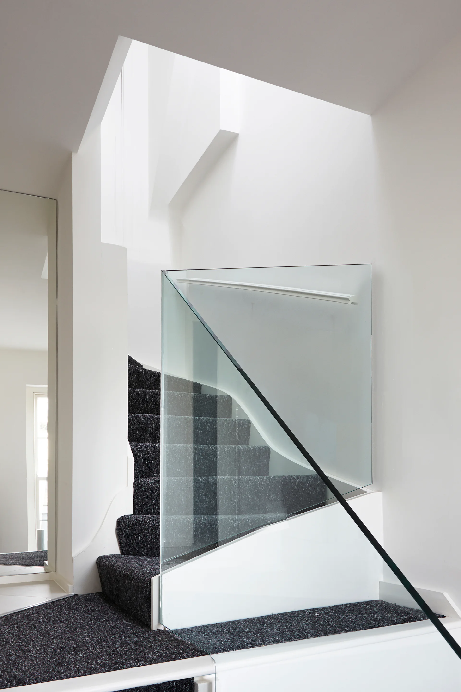 A minimalist modern staircase featuring charcoal grey carpeted steps, white walls, and a frameless glass balustrade under a bright skylight.