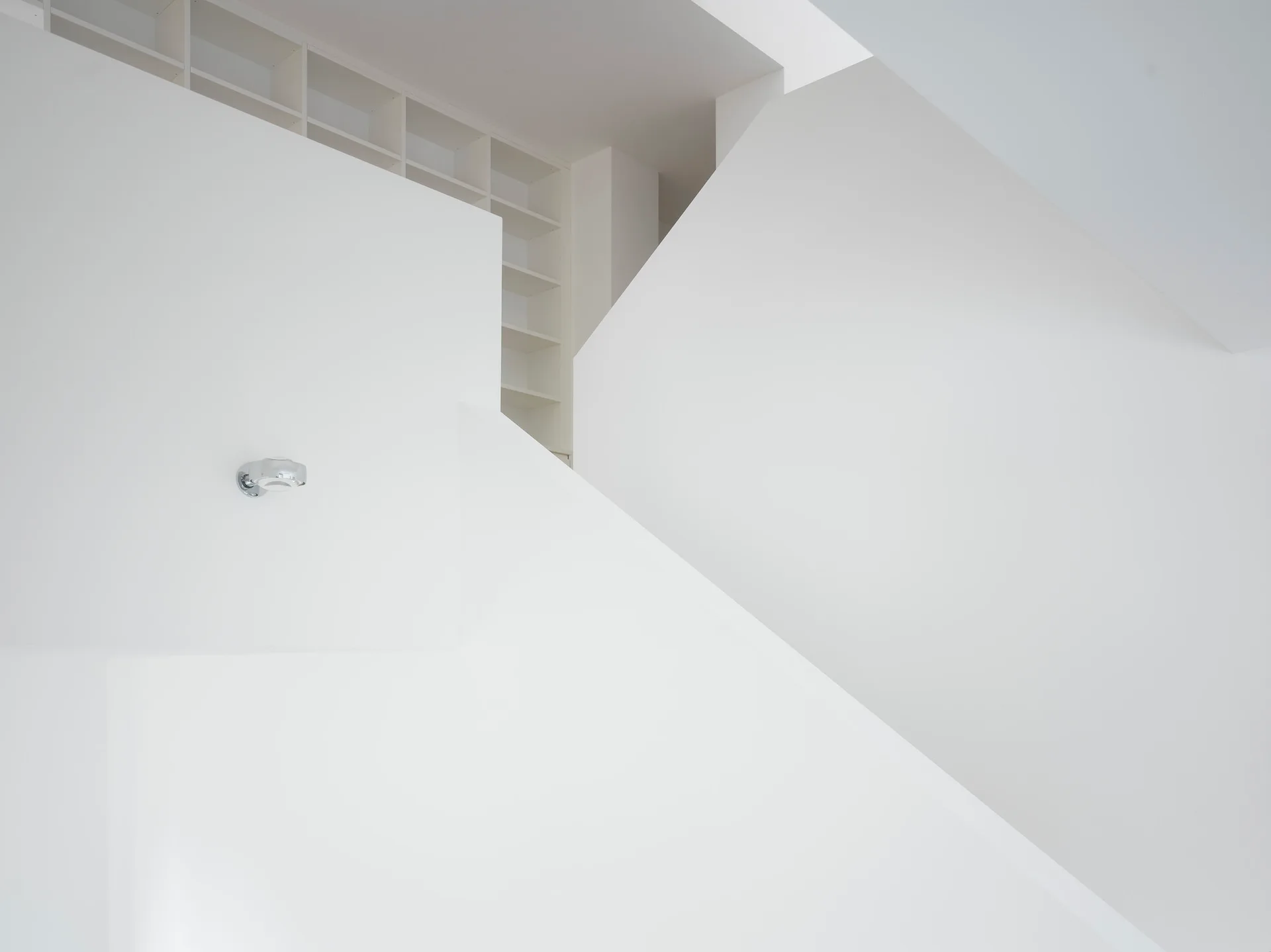 An angular white staircase with sharp geometric planes, a small silver wall sconce, and a white grid shelving unit in the background.