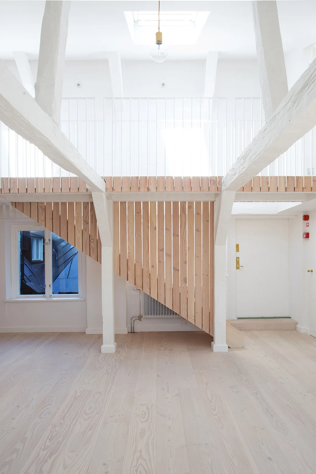 A minimalist white loft features a slatted light wood staircase beneath painted structural beams and a mezzanine with a white railing.