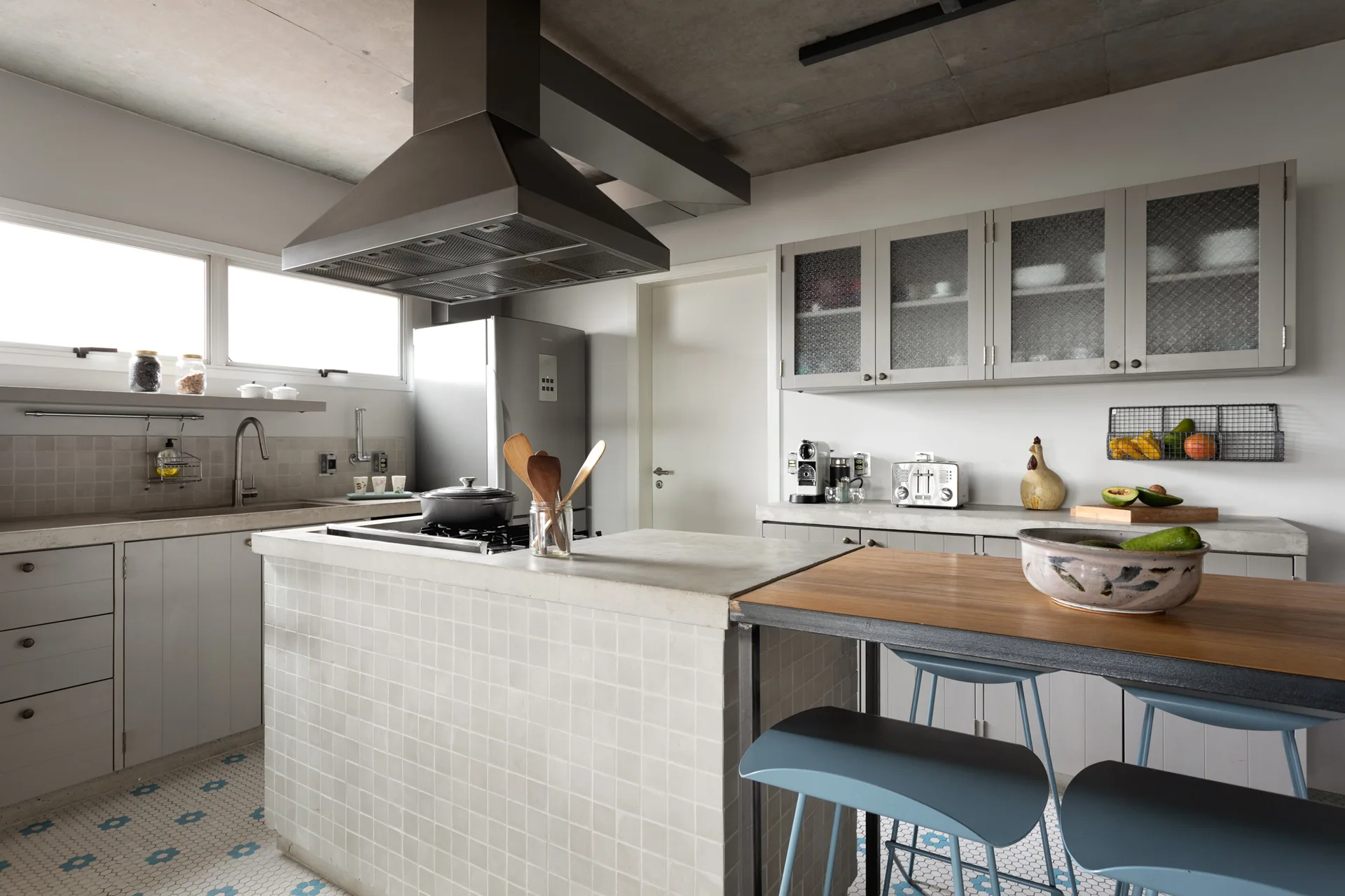 A modern kitchen featuring a central tiled island with a cooktop, blue bar stools, an exposed concrete ceiling, and patterned mosaic floor tiles.