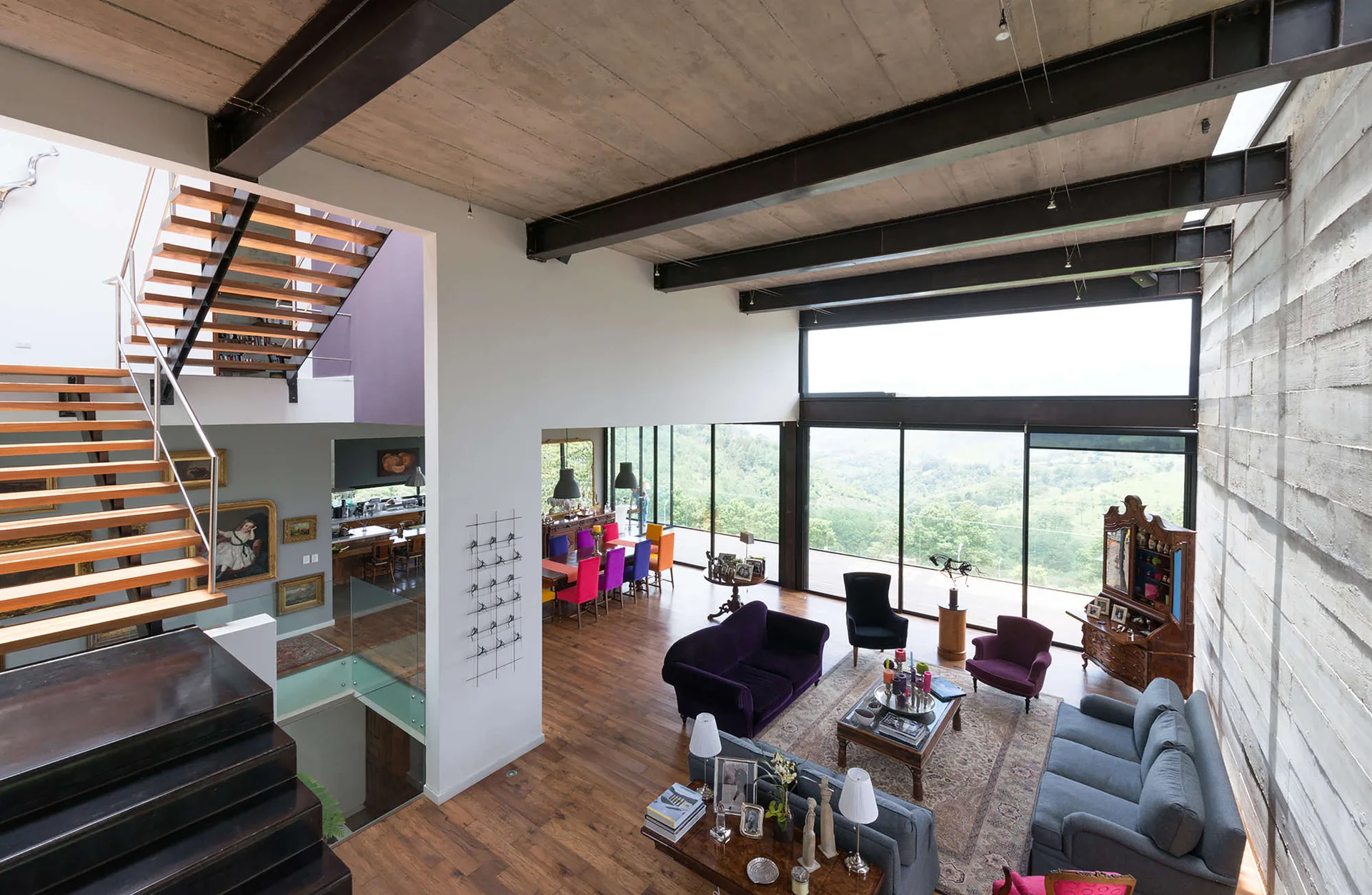 An industrial living room features a purple sofa, an open wooden staircase, exposed steel ceiling beams, and a large window overlooking a valley view.