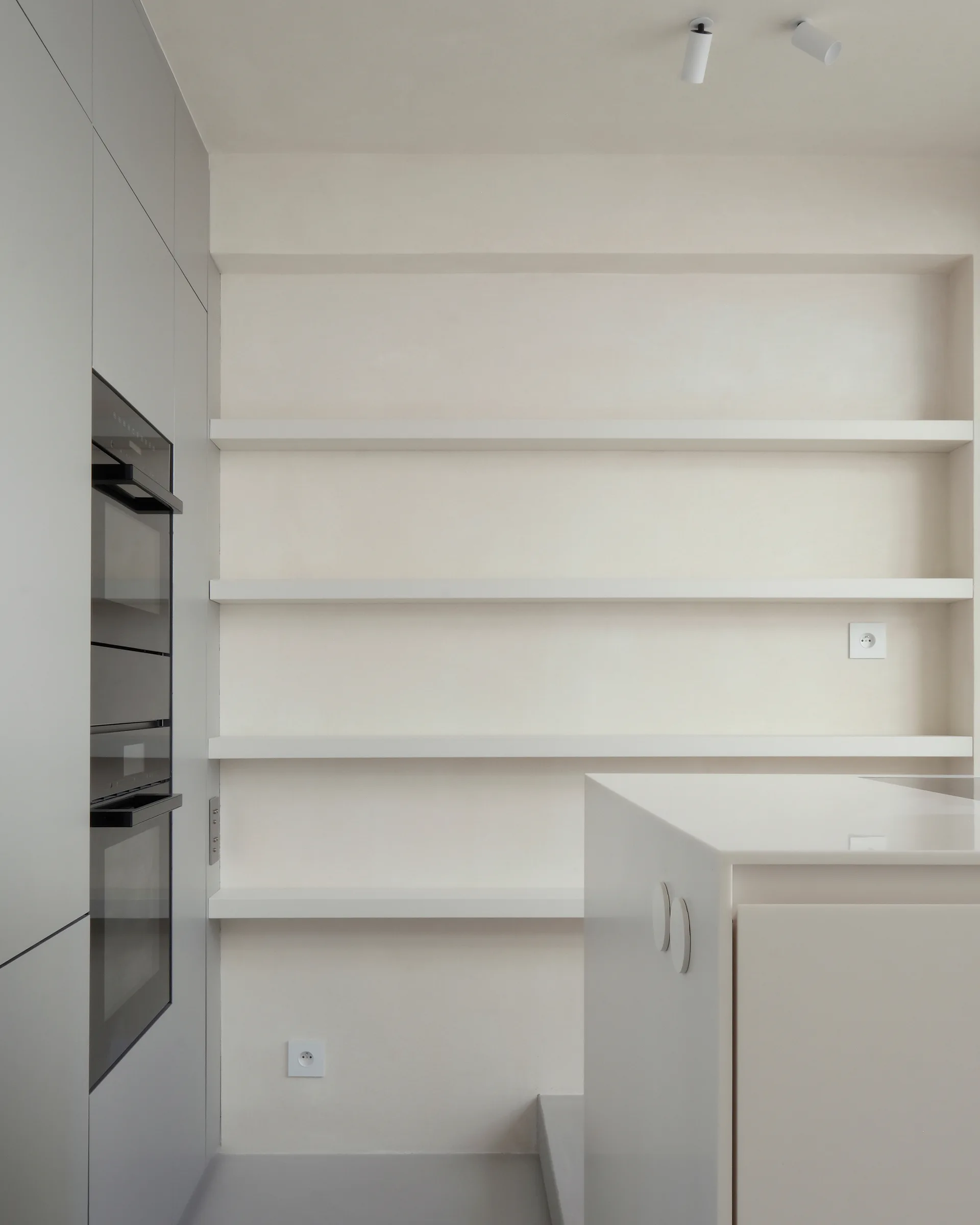 A minimalist kitchen features flat-front grey cabinetry with integrated ovens next to a wall niche with white open shelving.
