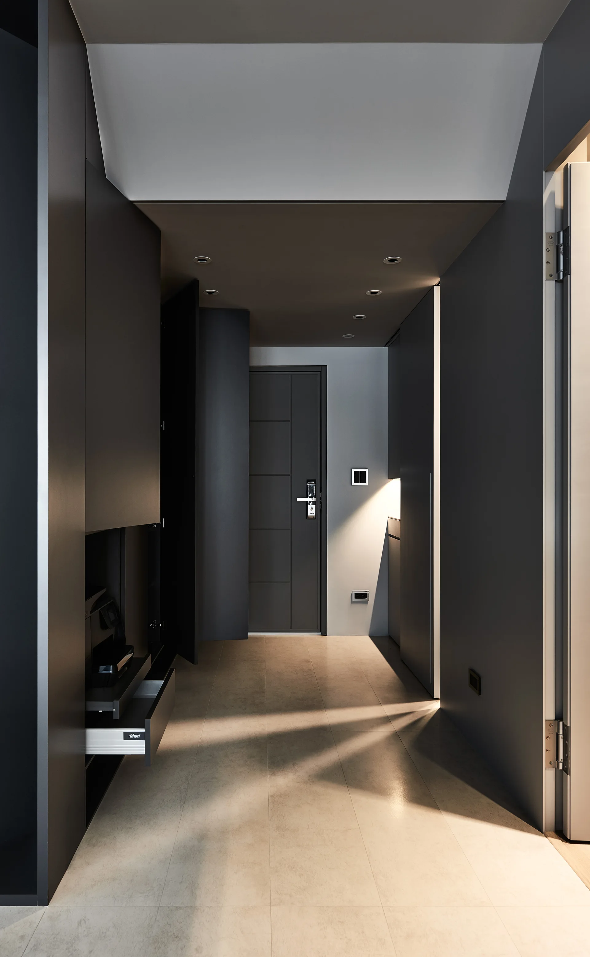 A minimalist hallway features floor-to-ceiling charcoal gray matte cabinetry, light beige floor tiles, and recessed ceiling spotlights.