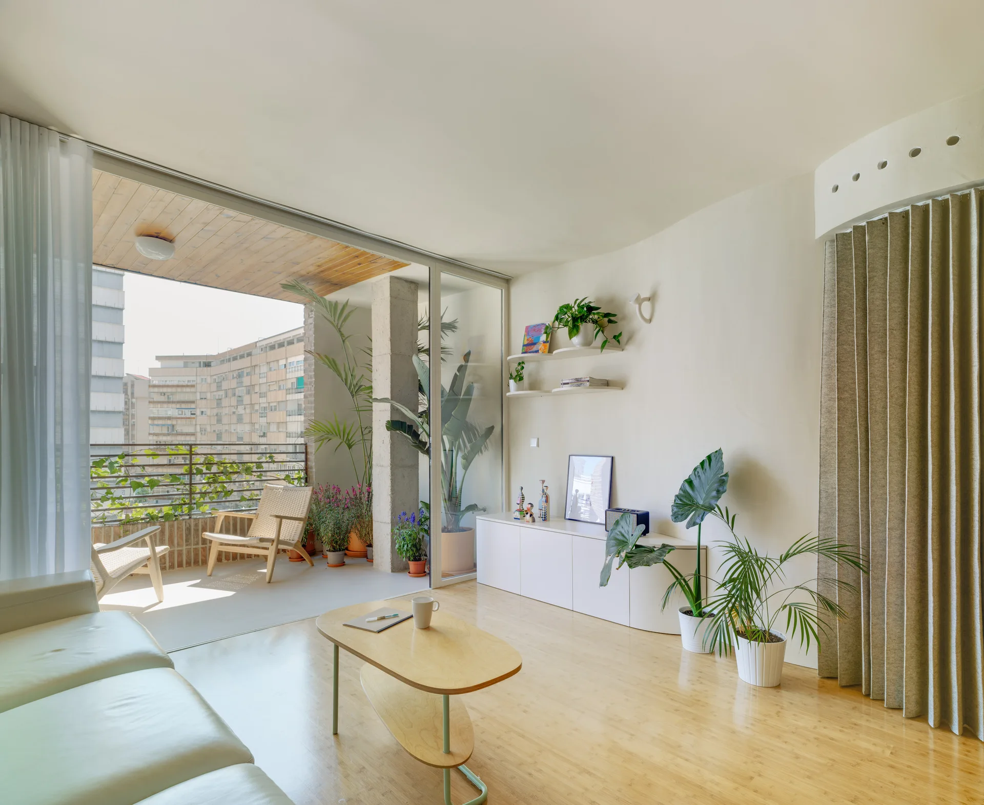 A minimalist living room with a white leather sofa and wood coffee table features sliding glass doors that open to a plant-filled outdoor balcony.