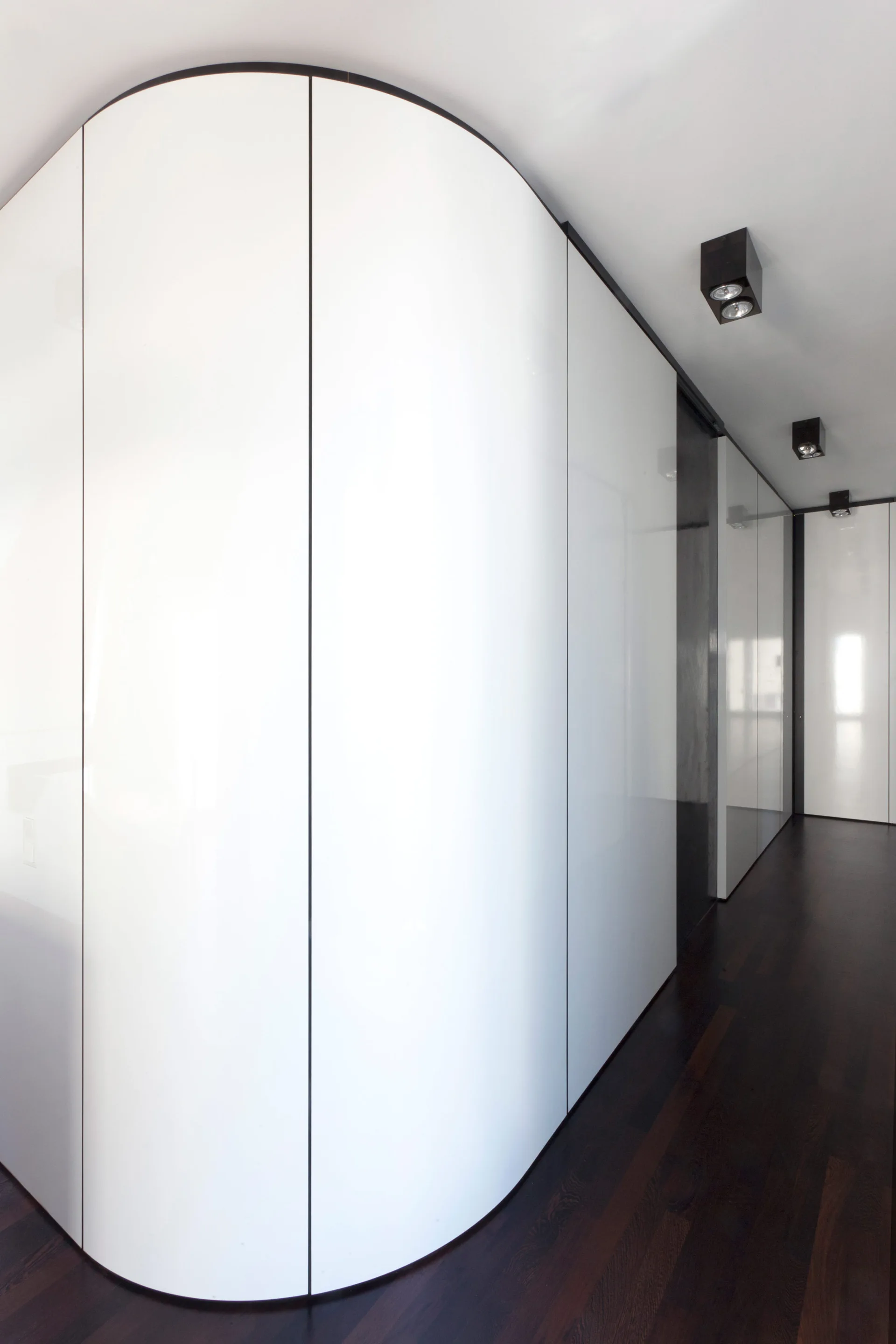 A minimalist hallway features a curved, glossy white paneled wall with thin vertical lines, dark wood flooring, and black ceiling spotlights.