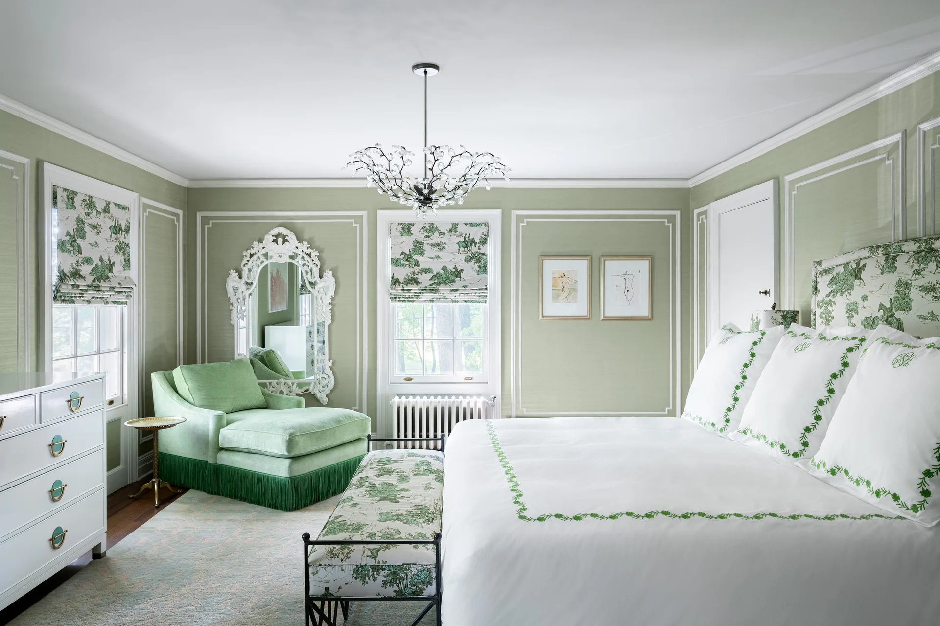 A traditional bedroom features sage green paneled walls, a white bed with green embroidered linens, and a matching fringed chaise longue.