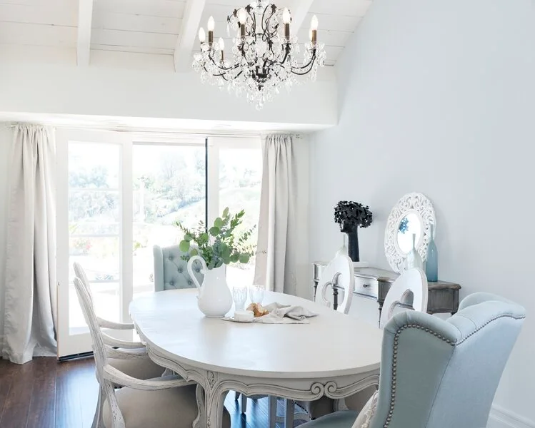 A bright dining room features an oval wooden table surrounded by light blue tufted chairs beneath a crystal chandelier and white beamed ceiling.