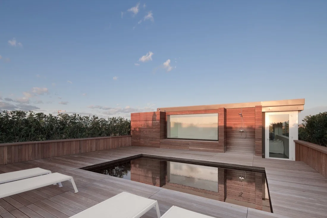 A rectangular swimming pool is inset into a weathered wood rooftop deck surrounded by matching wood-clad walls, white lounge chairs, and an outdoor shower.