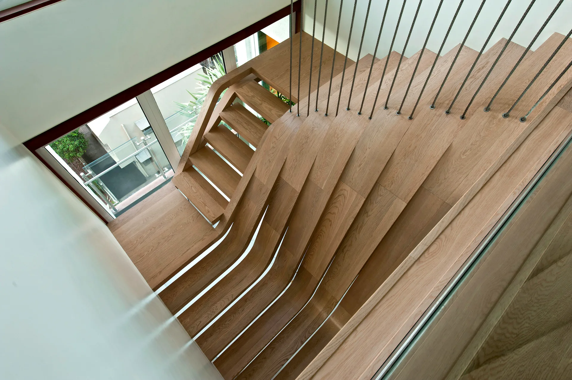 A high-angle view of a modern light wood staircase featuring curved wooden treads and vertical metal cable railings next to a white wall.