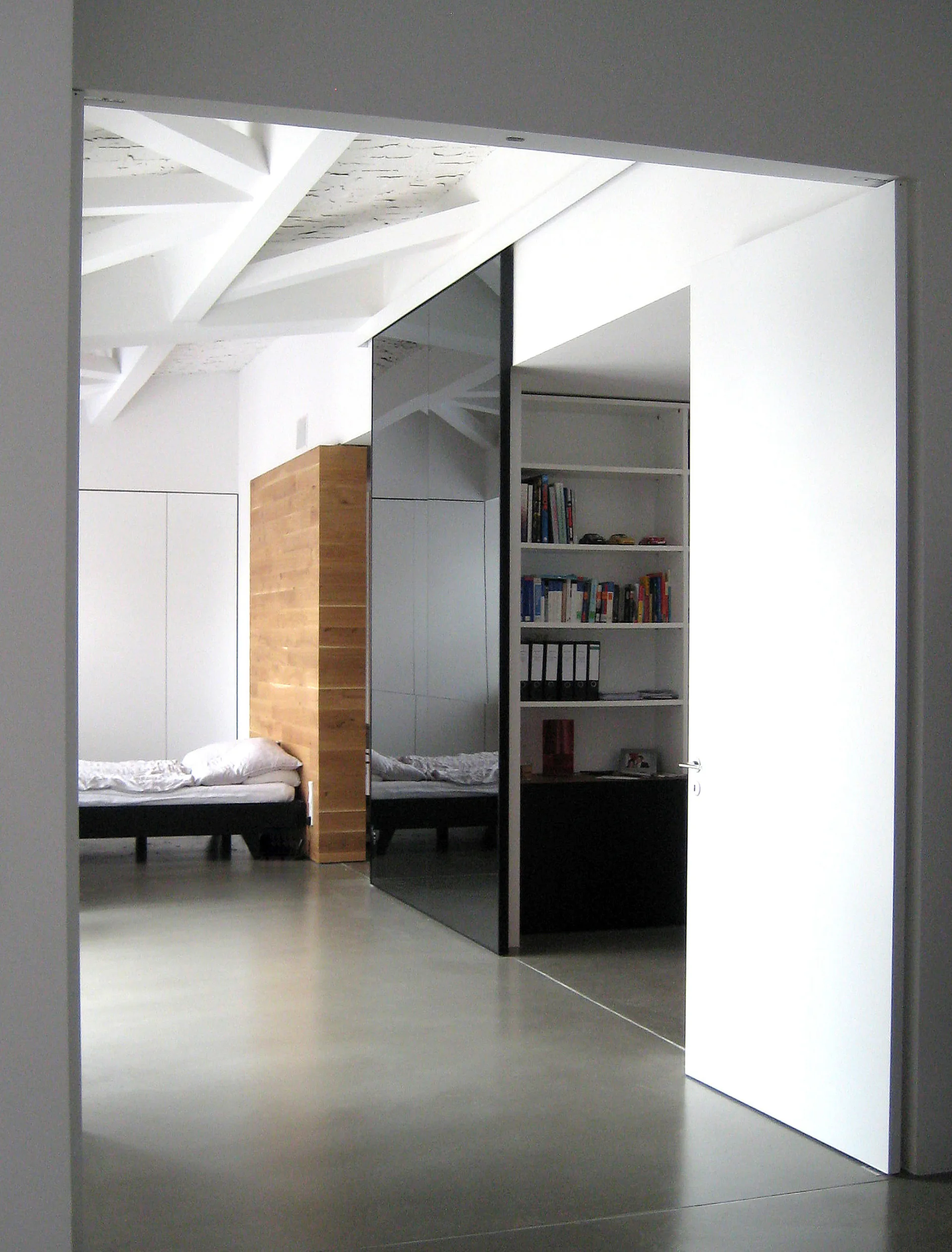 A minimalist bedroom seen through an open white door, featuring exposed white ceiling beams, a wood headboard, and a tall reflective black mirror panel.