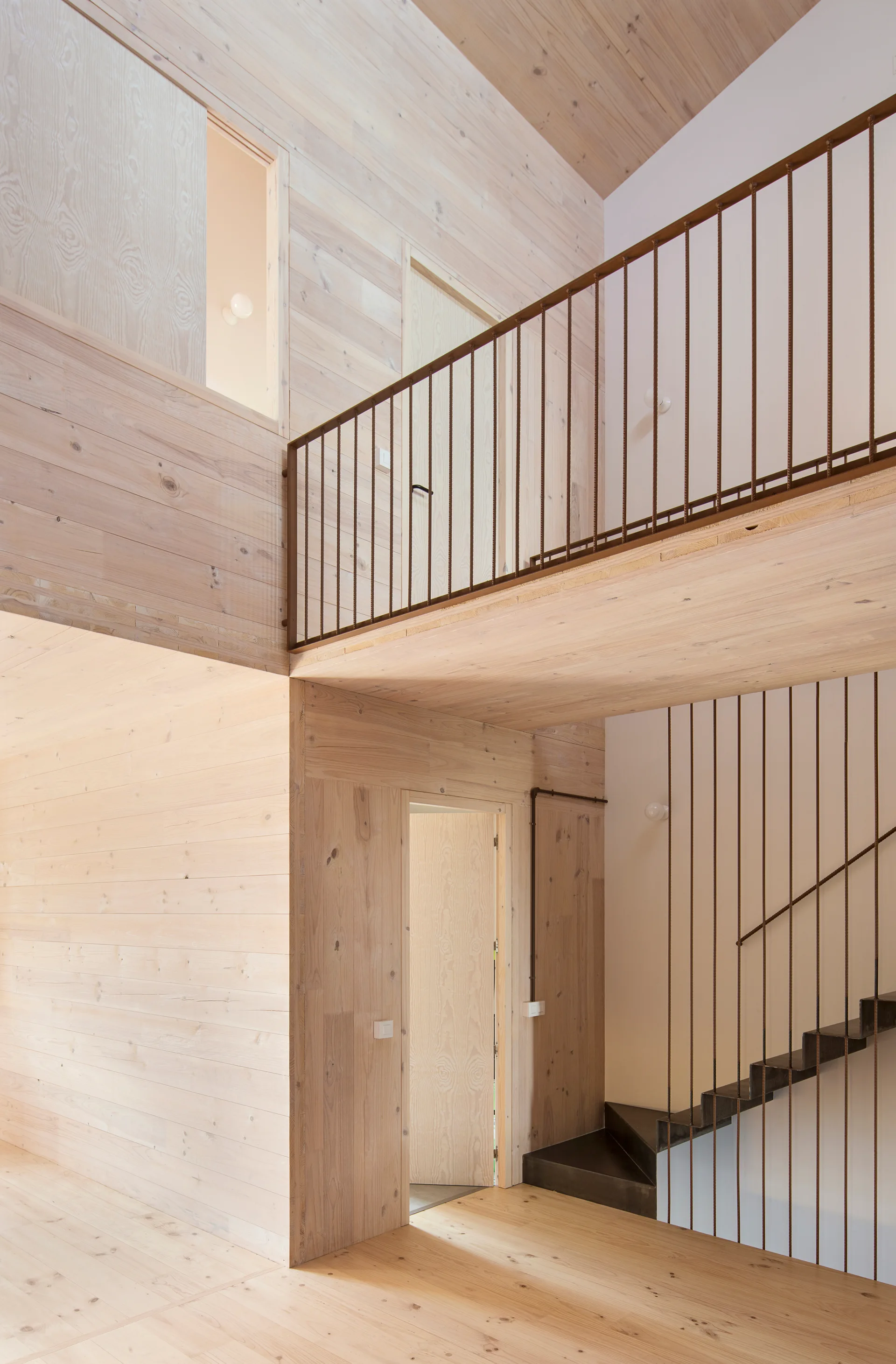 A modern multi-level hallway features extensive light wood paneling on walls and ceiling, a dark metal staircase, and thin vertical railings.