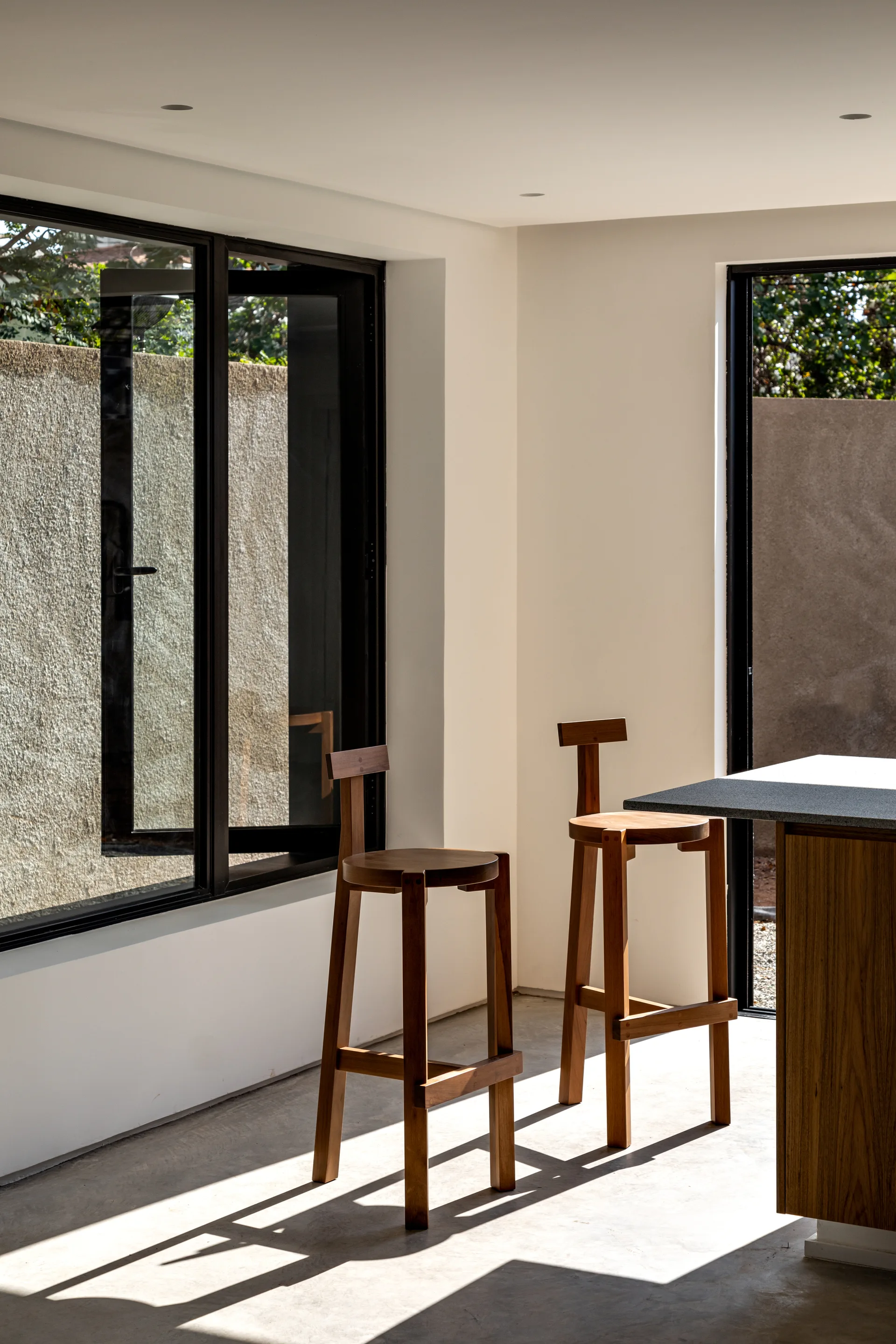 Two high-backed wooden bar stools stand by a kitchen island with a dark stone countertop in a room with black steel windows and concrete flooring.