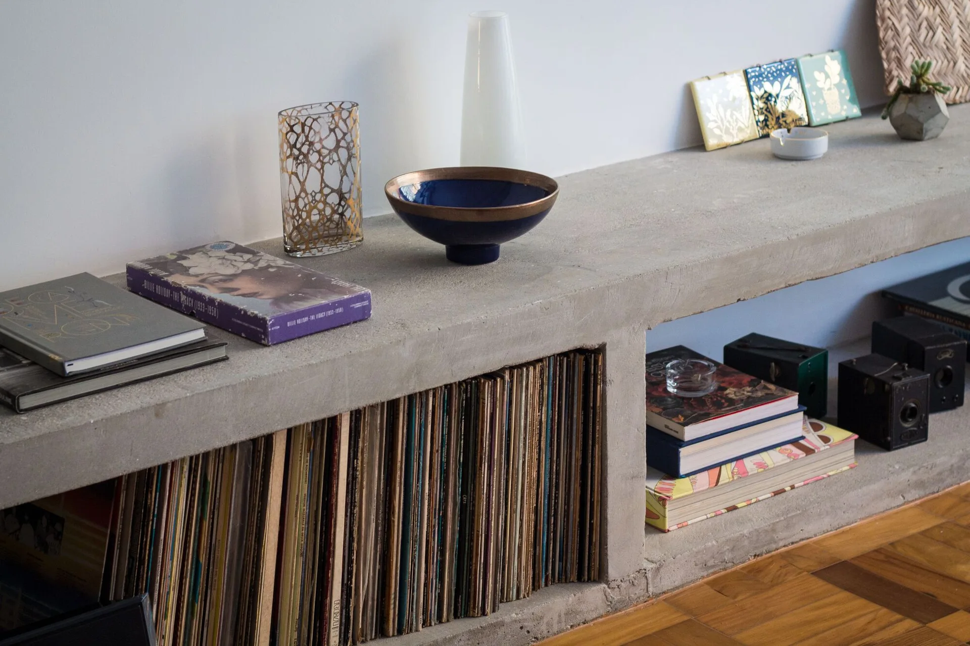 A low grey concrete shelving unit holding rows of vinyl records and books sits on a wooden parquet floor against a plain white wall.