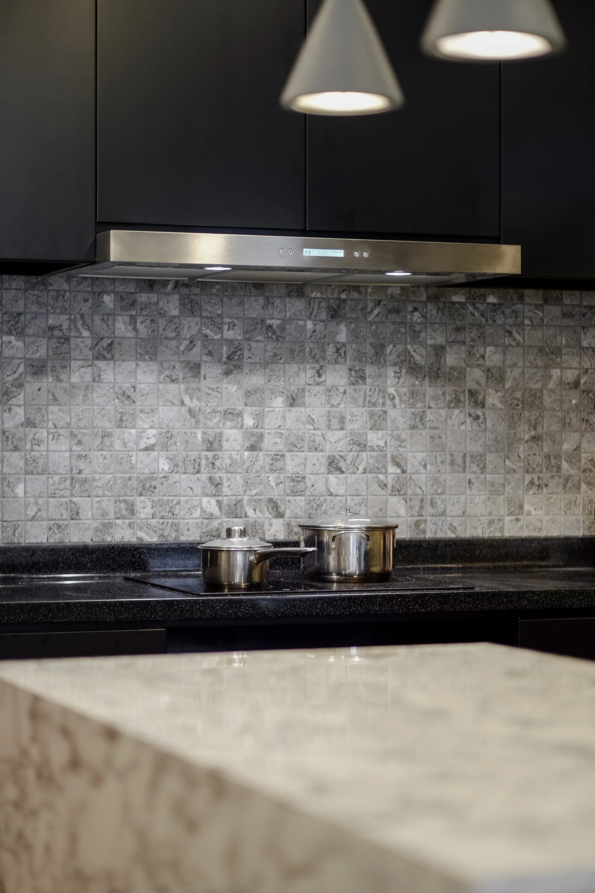 Stainless steel pots sit on a black cooktop beneath a sleek metal range hood, framed by matte black cabinets and a grey stone mosaic tile backsplash.