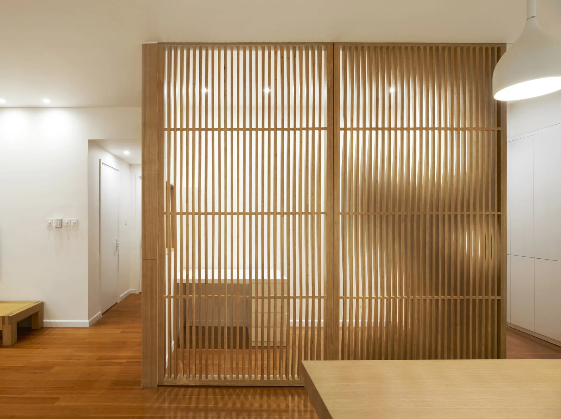 A minimalist room featuring a vertical wooden slat screen partition, polished wood flooring, and clean white walls.