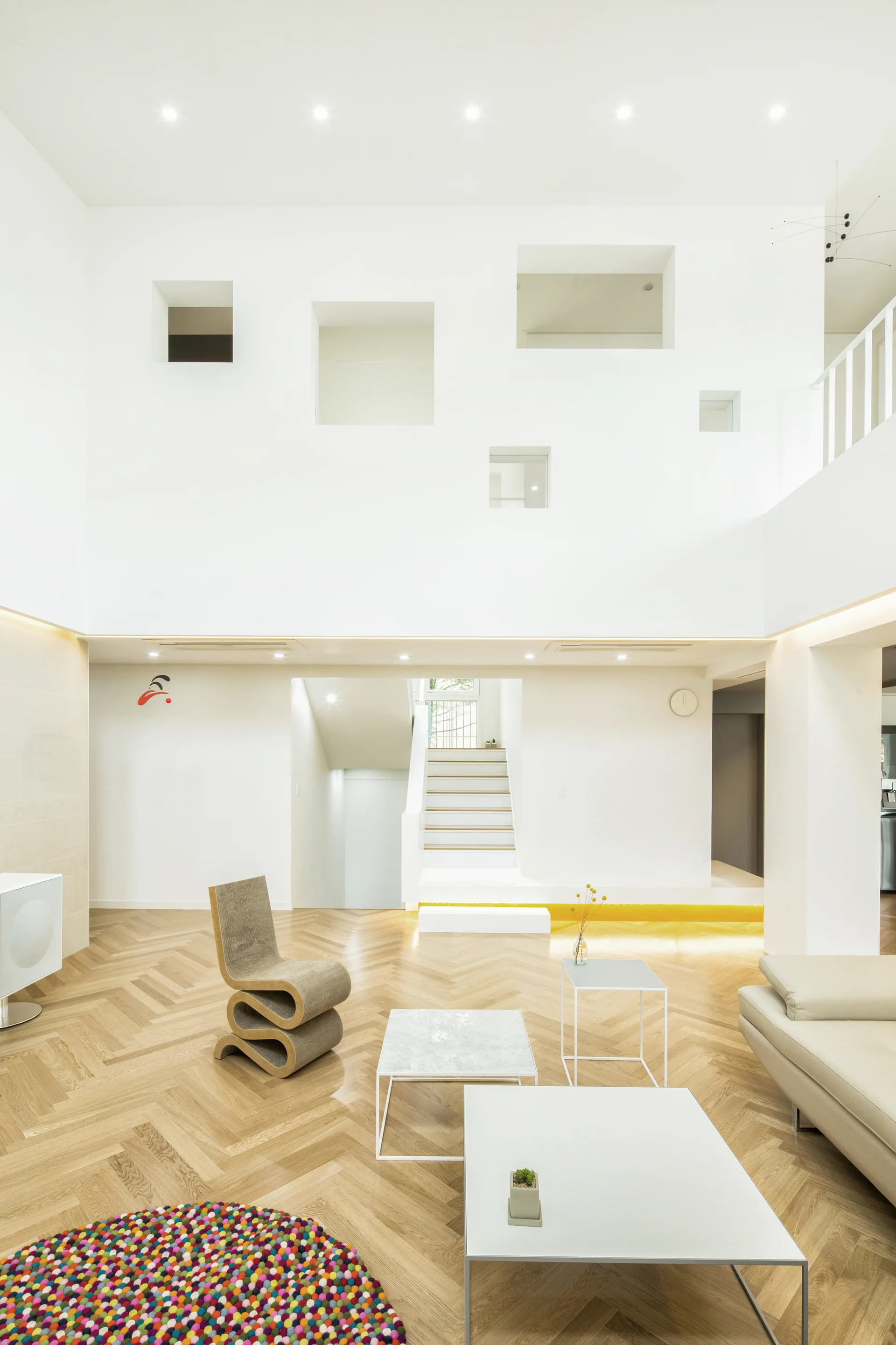A bright minimalist living room features light wood herringbone flooring, a wavy cardboard accent chair, and white walls with square architectural cutouts.