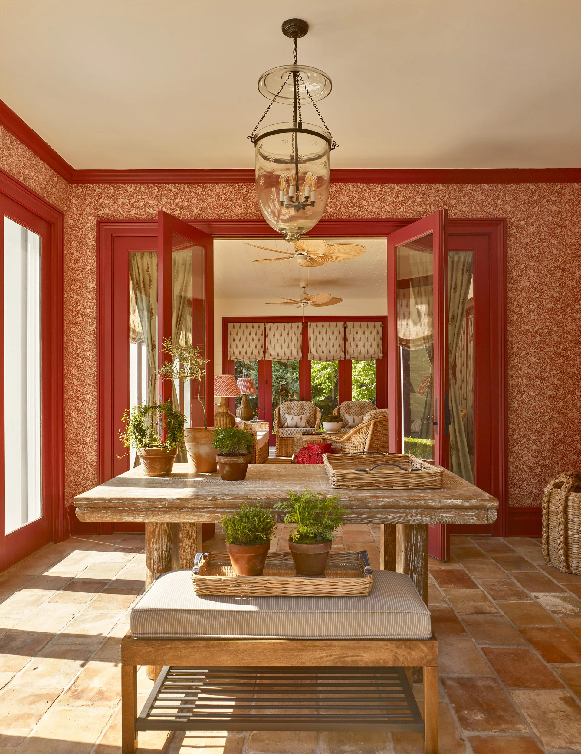 A distressed wood table sits on a terracotta tile floor in a room featuring red painted French doors and botanical patterned wallpaper.