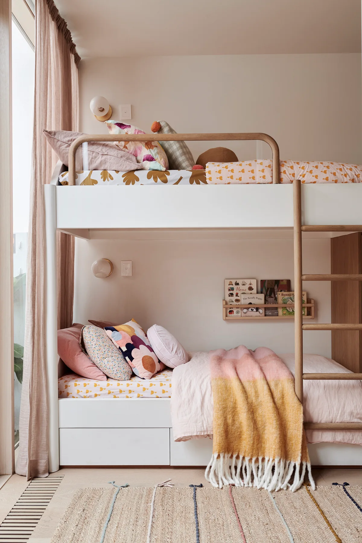 A white wooden bunk bed with a natural wood ladder and safety rails, decorated with soft pink and yellow textiles in a light-filled kids' bedroom.