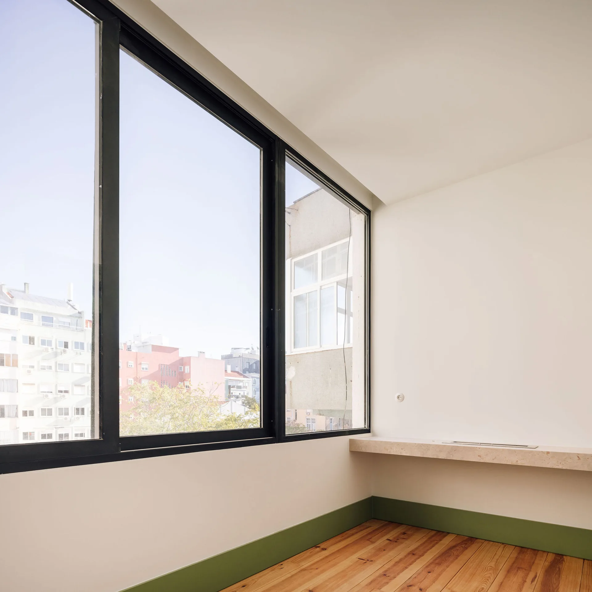 An empty room featuring light wood floors, a green baseboard, white walls, large black-framed windows, and a floating stone ledge.