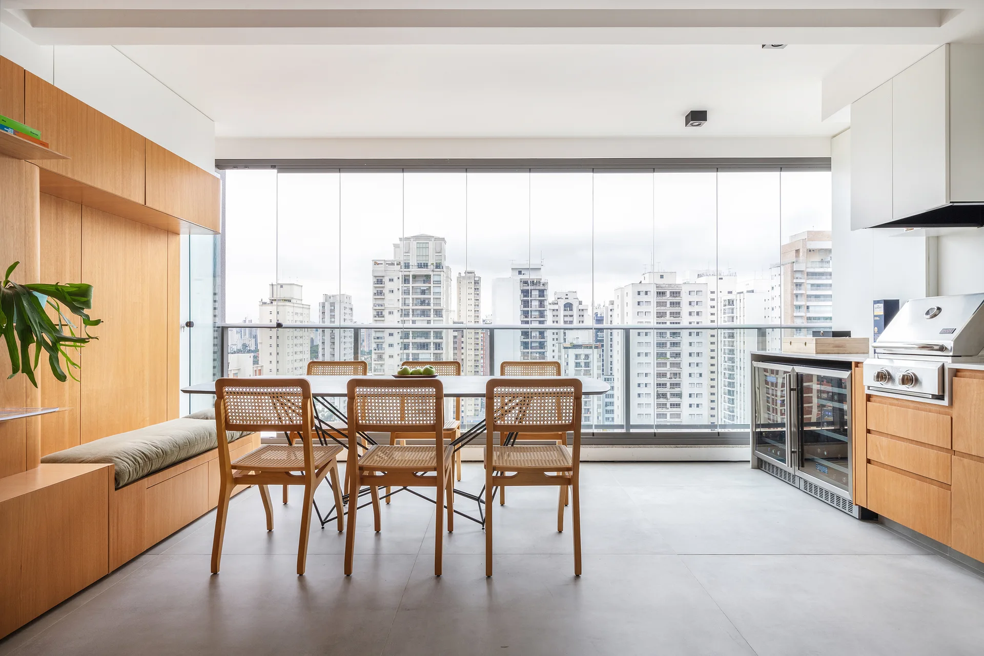 A modern dining table with cane-back chairs sits inside a glass-enclosed apartment balcony featuring wooden built-ins and views of city high-rises.