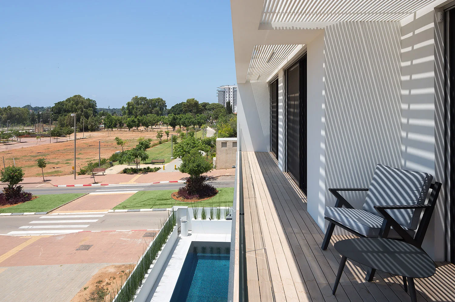 A modern wood-planked balcony with a black metal armchair and table features a slatted overhead pergola and a glass railing overlooking a pool.