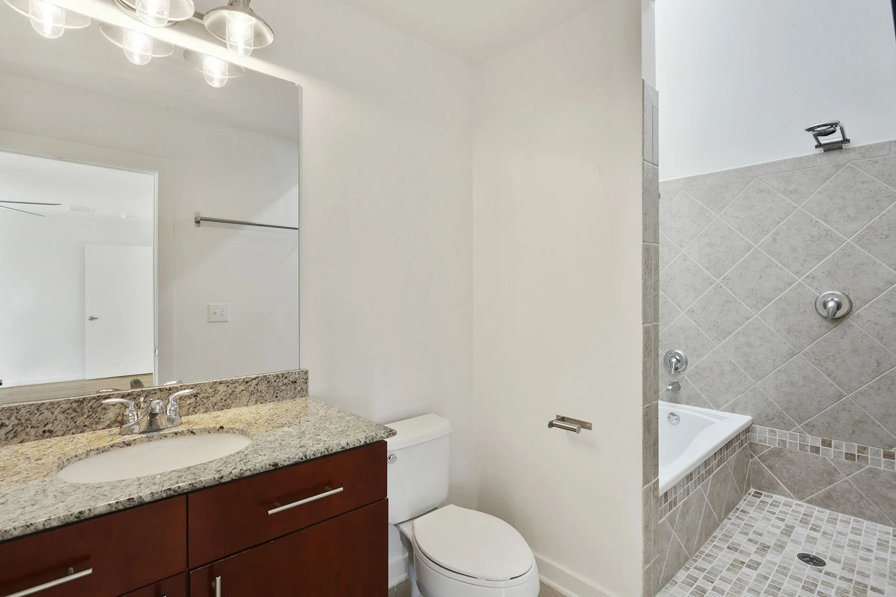 A modern bathroom features a dark wood vanity with a speckled stone countertop, a white toilet, and a tiled shower with a bathtub.